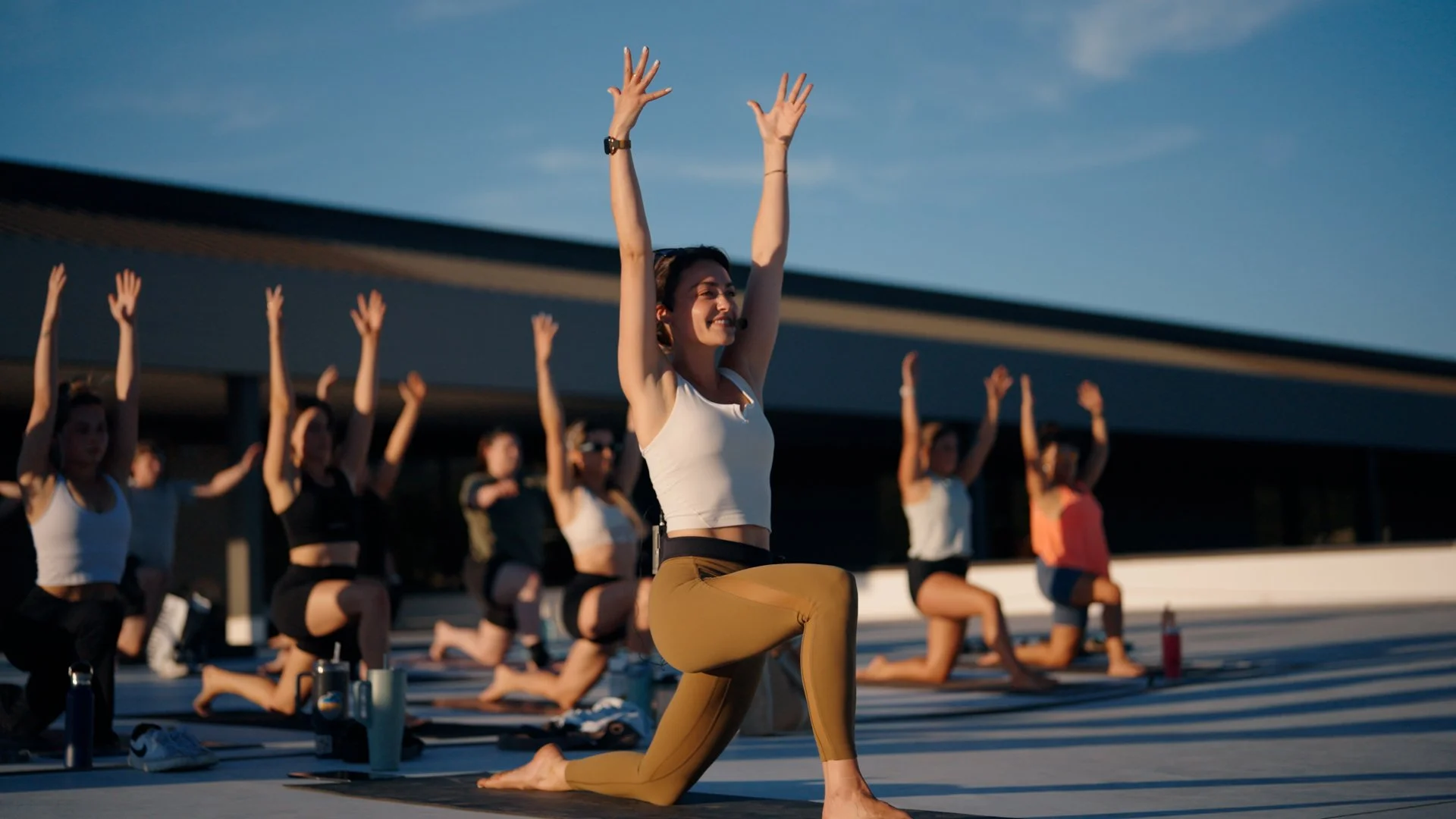 Group of women practicing yoga outdoors during sunset, with one woman in foreground performing a kneeling pose with arms raised.