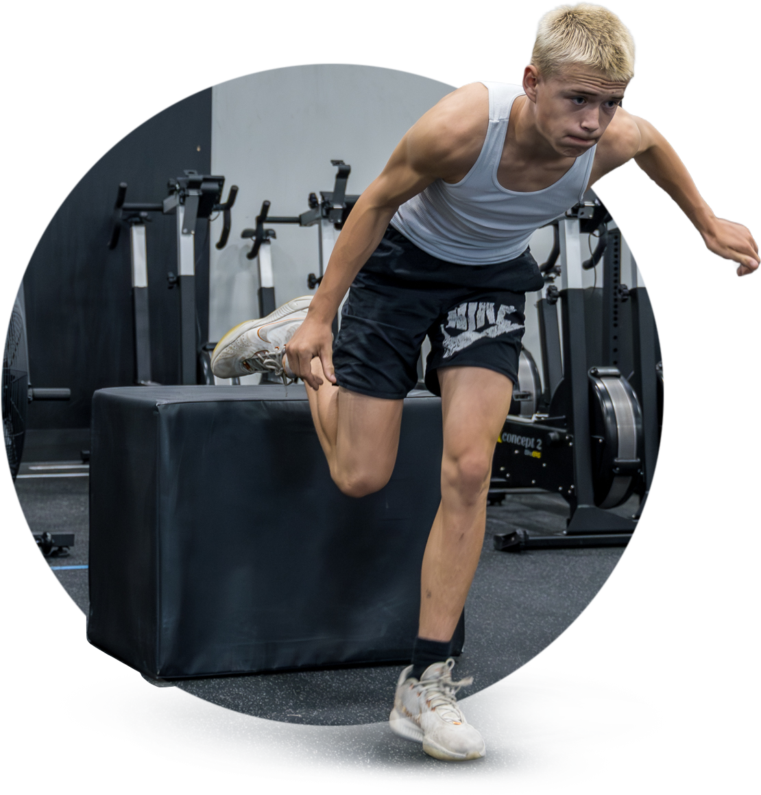 Young man with blonde hair wearing a gray tank top and black shorts doing a step-up exercise on a black plyometric box in a gym.