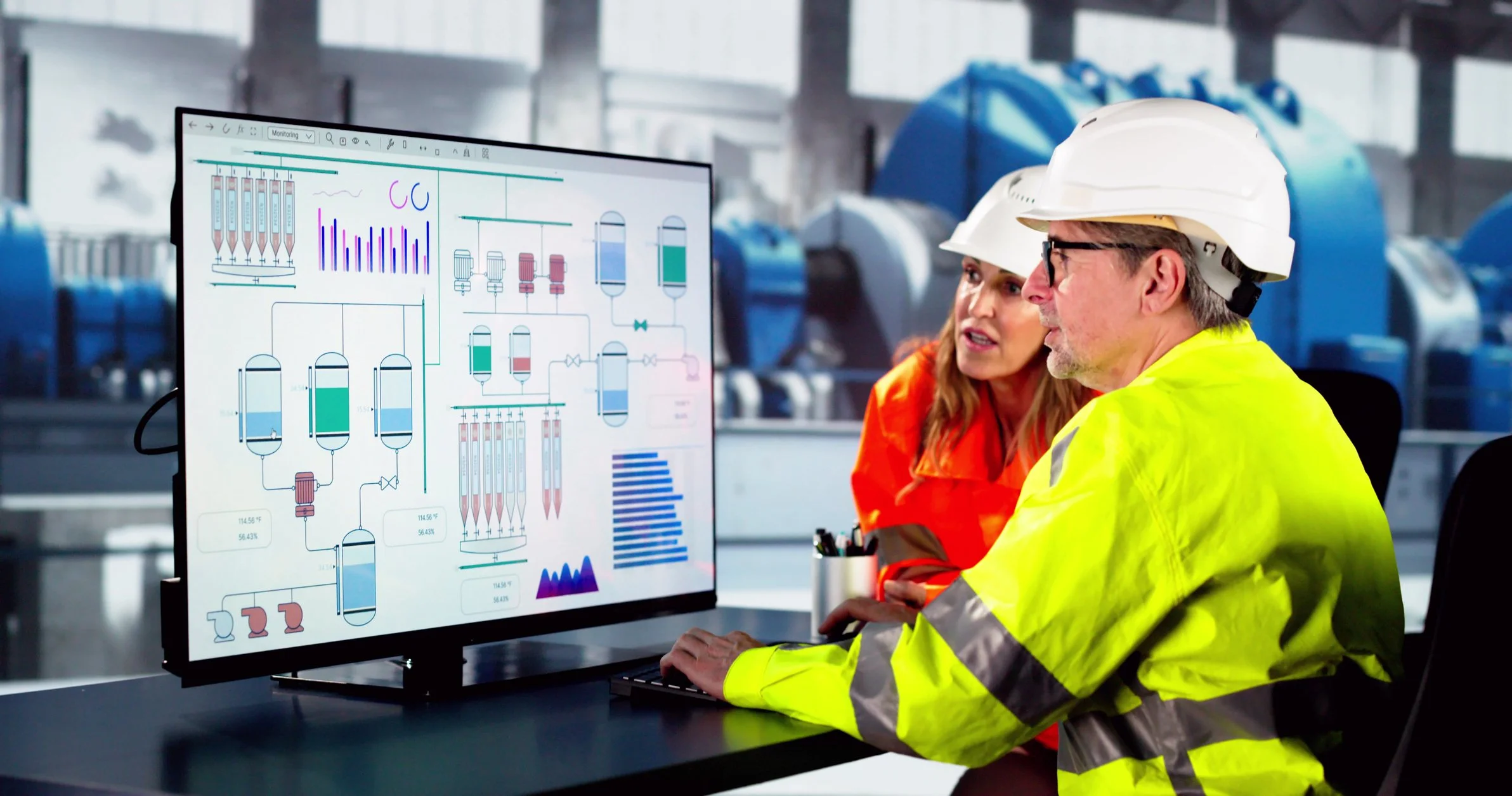 Two engineers in a factory, wearing safety helmets and high-visibility jackets, discussing a digital interface displayed on a monitor, which shows technical schematics and data.
