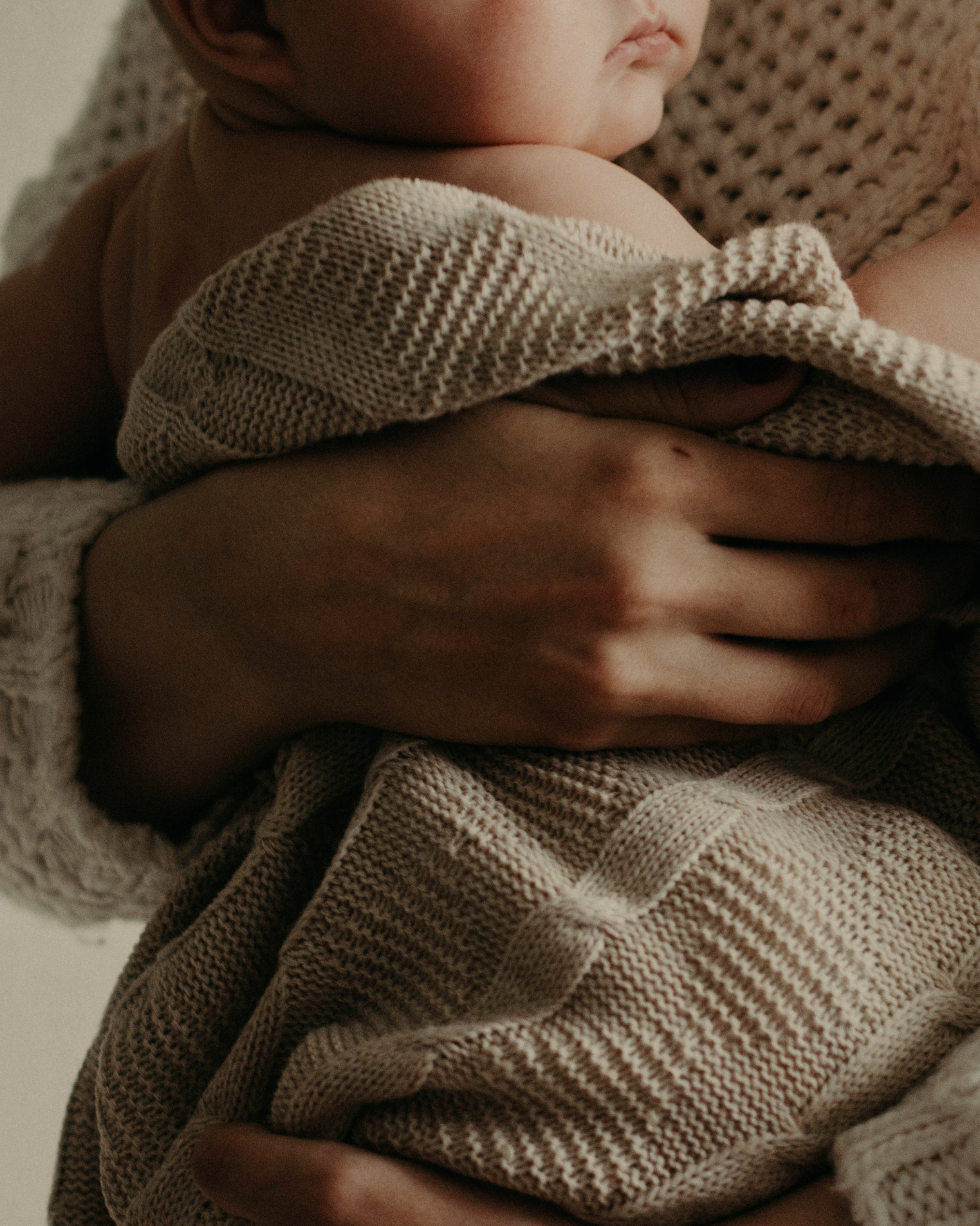 Close-up of a baby being held, with focus on the baby's shoulder, part of the face, and hand. The baby wears a knitted beige and brown striped sweater, and is held by an adult with a hand visible supporting the baby's body.