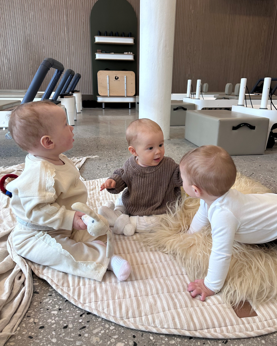 Close-up of a baby being held, with focus on the baby's shoulder, part of the face, and hand. The baby wears a knitted beige and brown striped sweater, and is held by an adult with a hand visible supporting the baby's body.