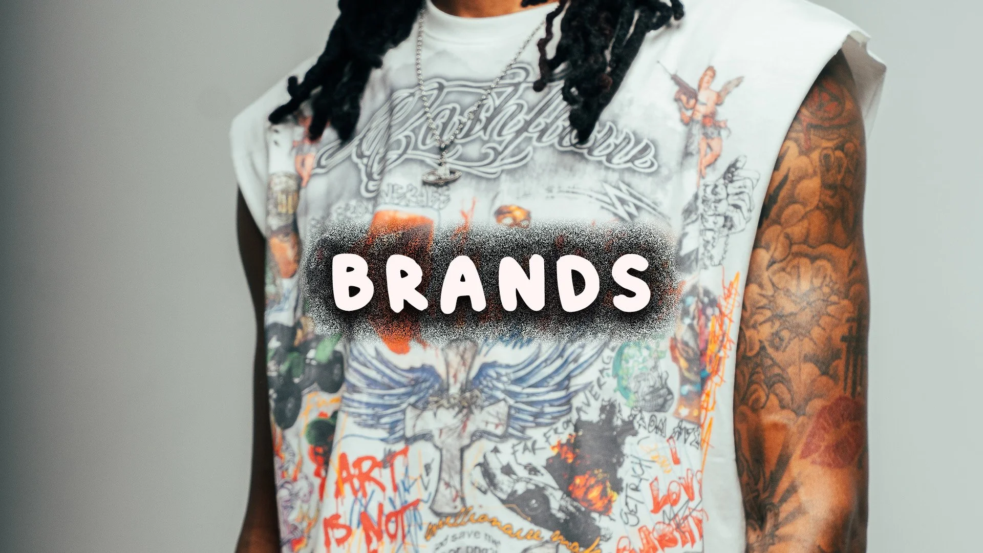 Close-up of a person wearing a graphic sleeveless shirt with various illustrations and text. The person has long, dark dreadlocks and visible tattoos on their arm. The word 'BRANDS' is spray-painted on the shirt in white letters over a black spray-pa