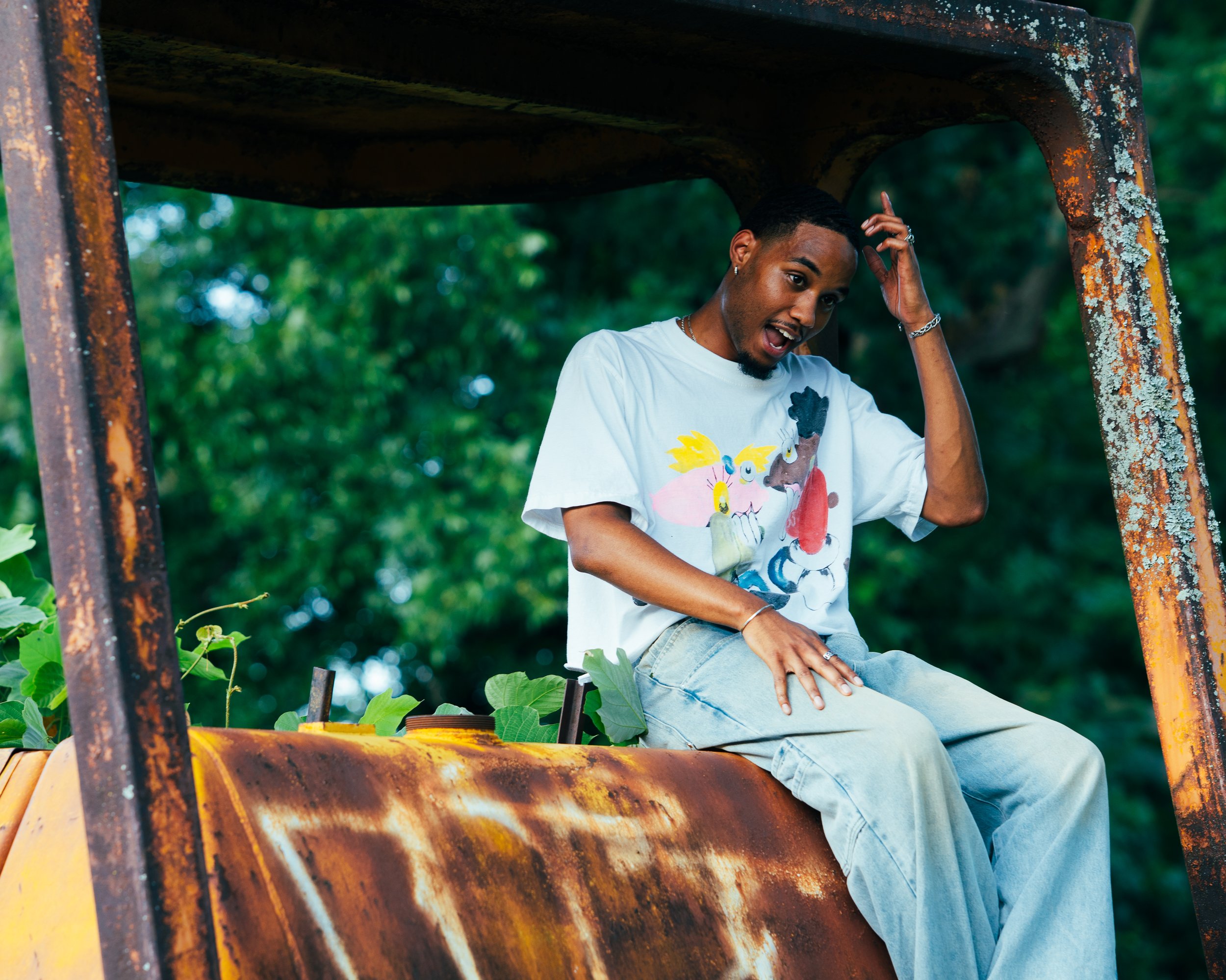 A young man with dark hair and a beard sitting on an old, rusty yellow tractor, wearing a white T-shirt with colorful animated characters and light blue jeans, outdoors with green trees in the background.