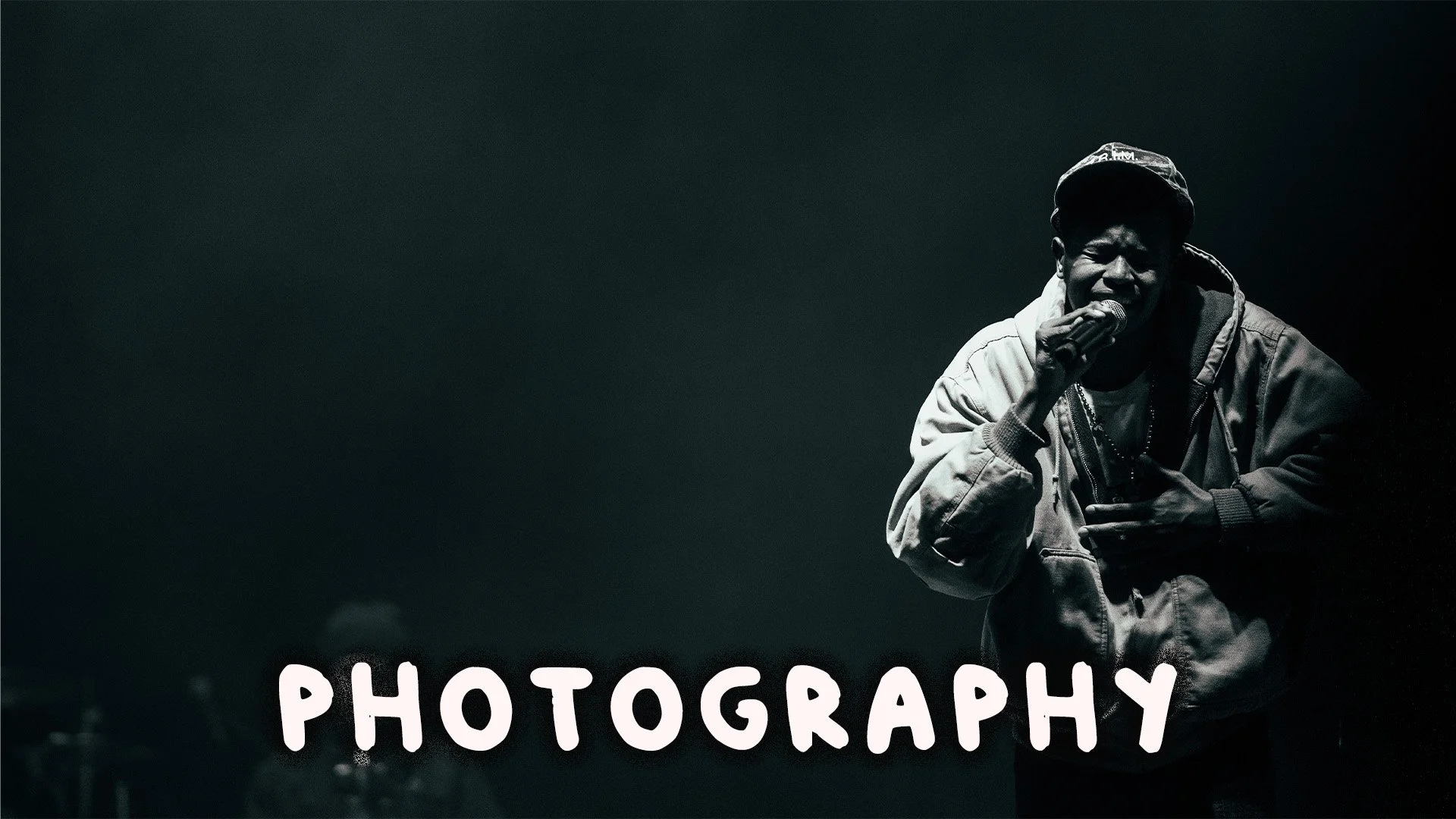 A black-and-white photo of a man singing into a microphone, wearing a jacket and cap, with the word 'PHOTOGRAPHY' in stylized font at the bottom.