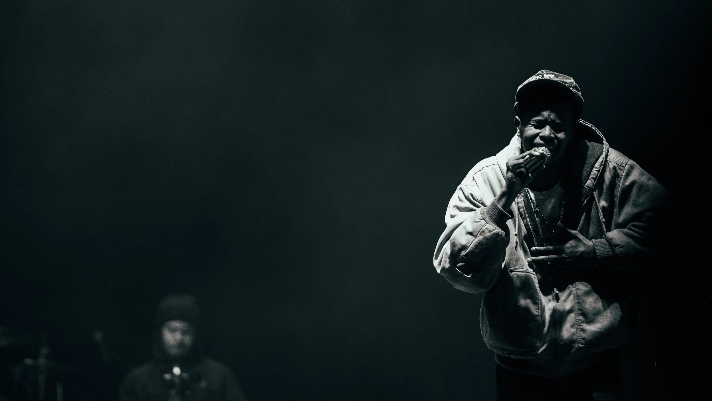 A man singing passionately into a microphone on stage, wearing a jacket and cap, with a dark background and another person in the background.