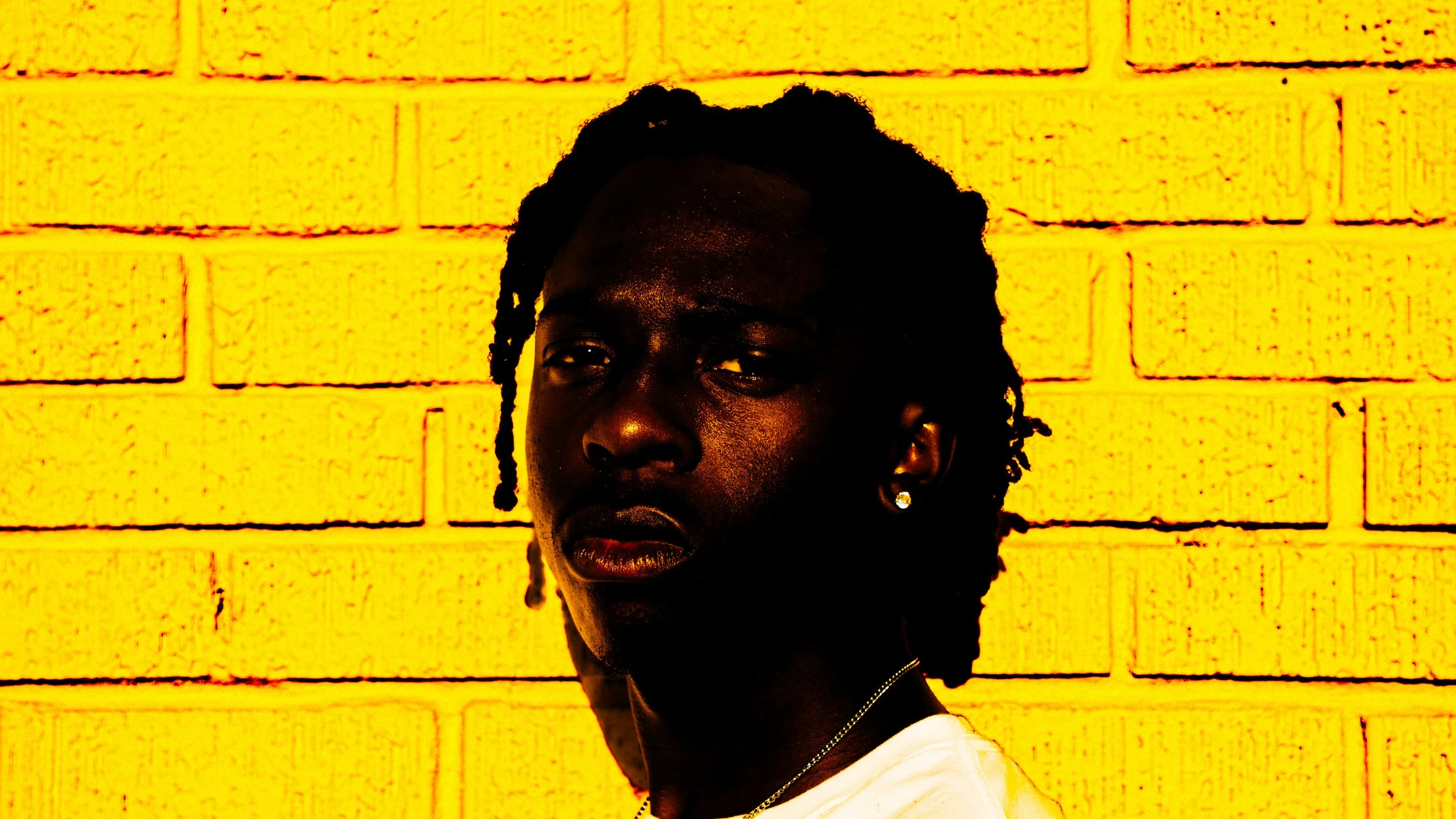 Portrait of a young Black man with braided hair, wearing earrings and a white shirt, standing against a yellow brick wall.