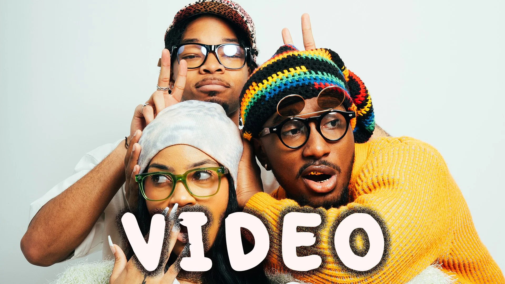 Three young adults posing playfully against a plain background, with the word 'VIDEO' written in white and black text in front of them.