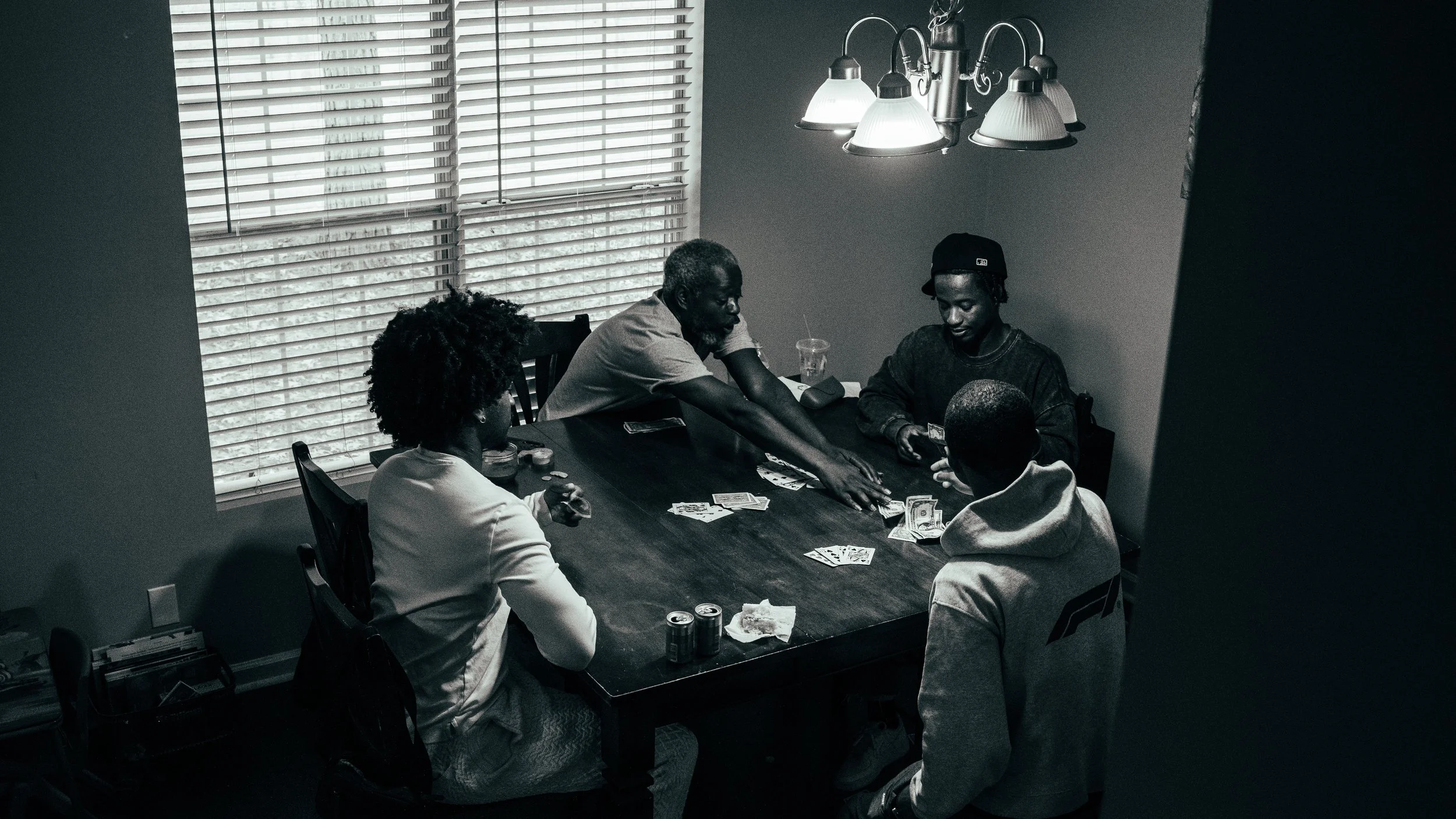 Four people are sitting around a dining table playing cards. The room has large windows with blinds, and a hanging light fixture illuminates the scene. One person is standing, reaching across the table.