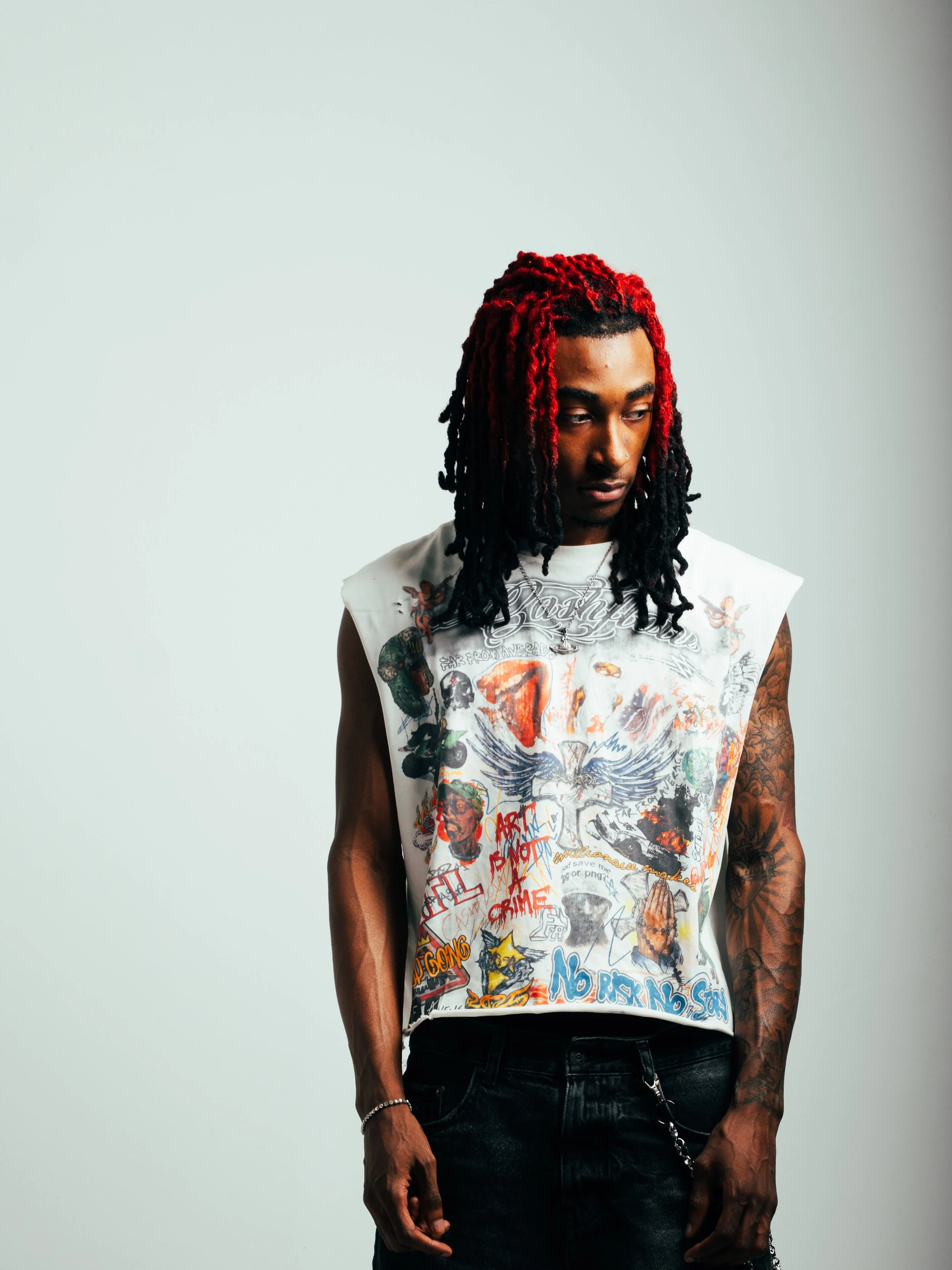 A young man with red and black dreadlocks wearing a sleeveless graphic T-shirt and black jeans standing against a plain light background, looking down.