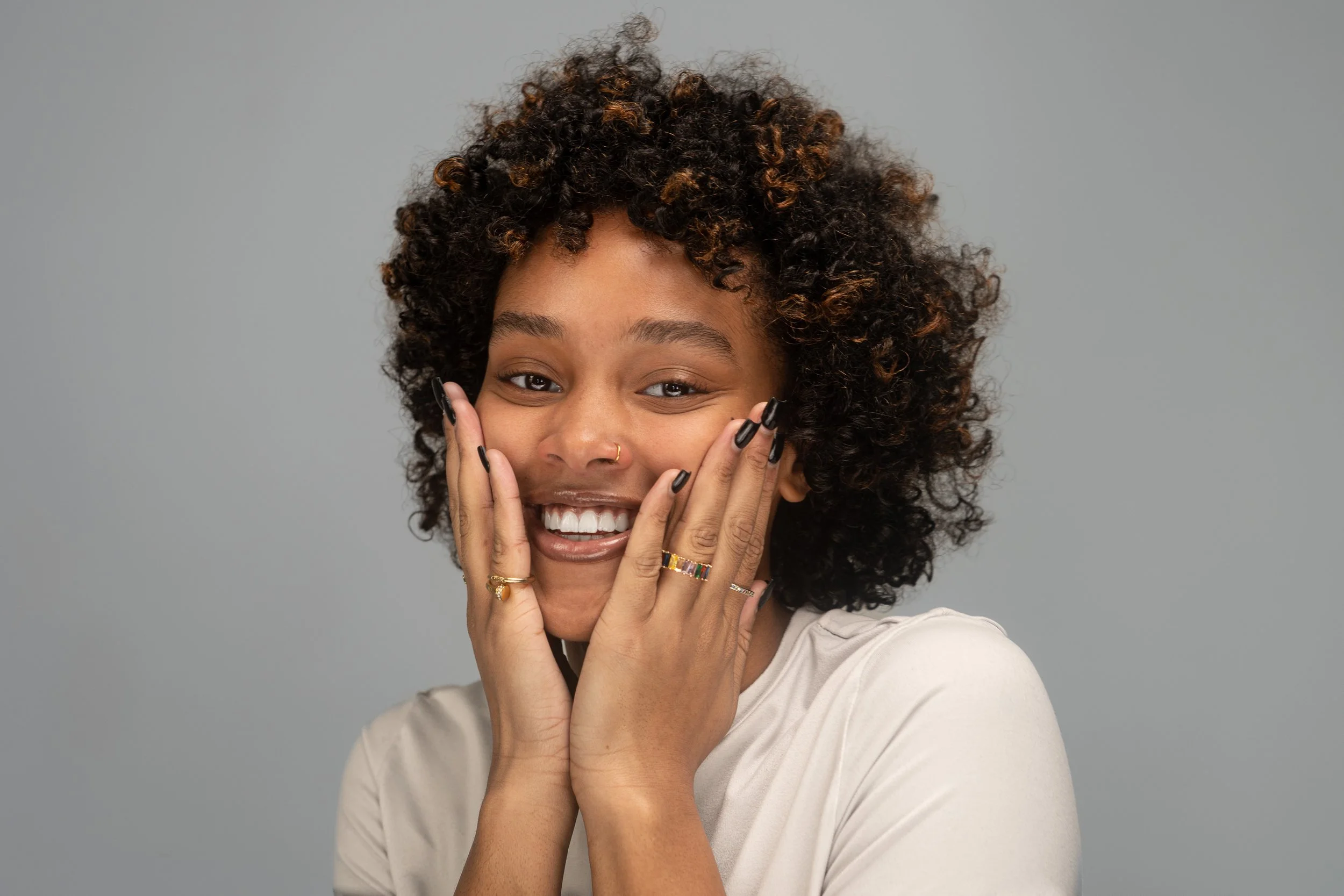 A young woman with curly hair, smiling and holding her face with her hands, wearing rings and rings on her fingers, against a plain gray background.