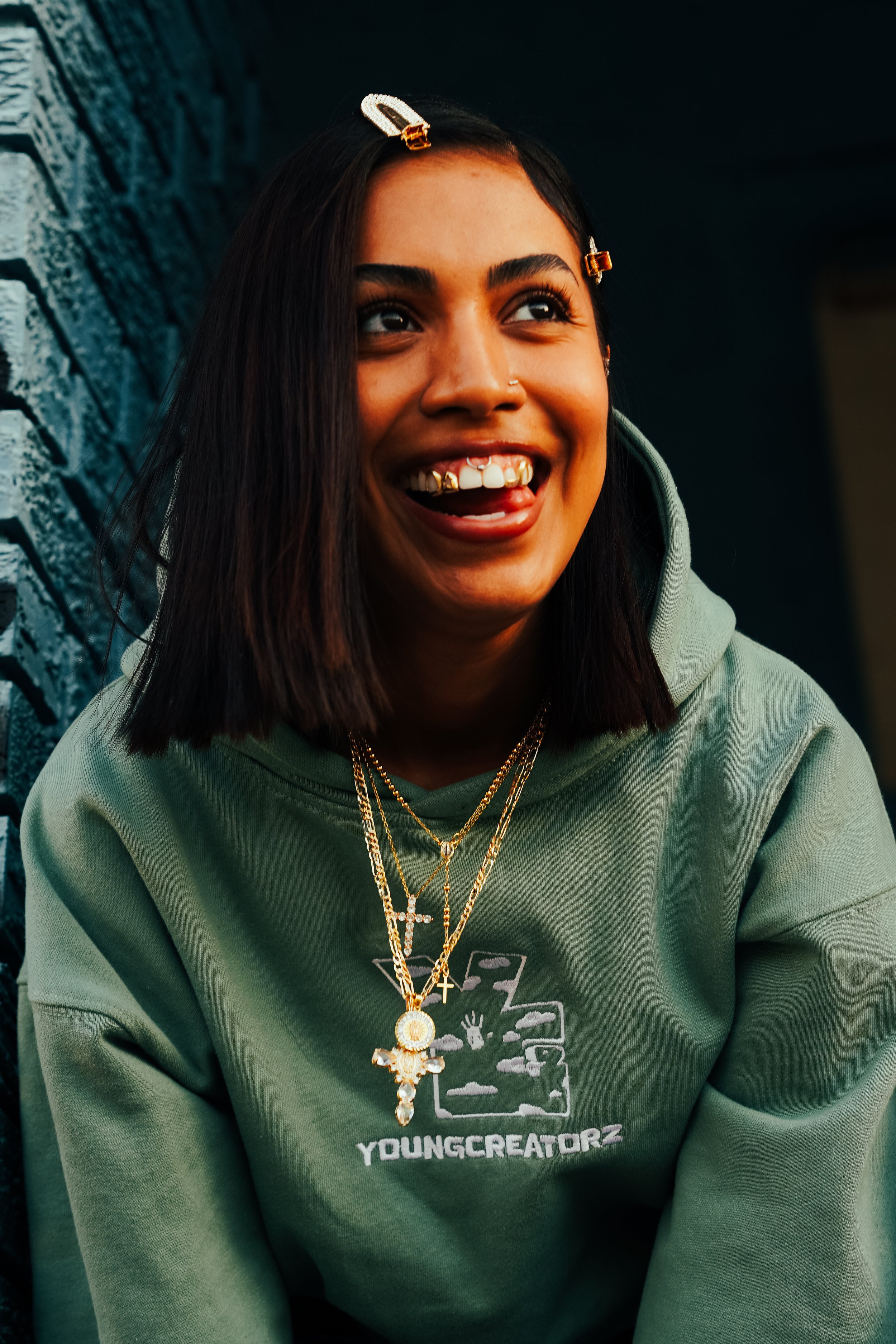 A woman with medium dark hair, wearing layered gold chains and a green hoodie with 'YOUNGCREATORZ' logo, smiling playfully with her tongue out.