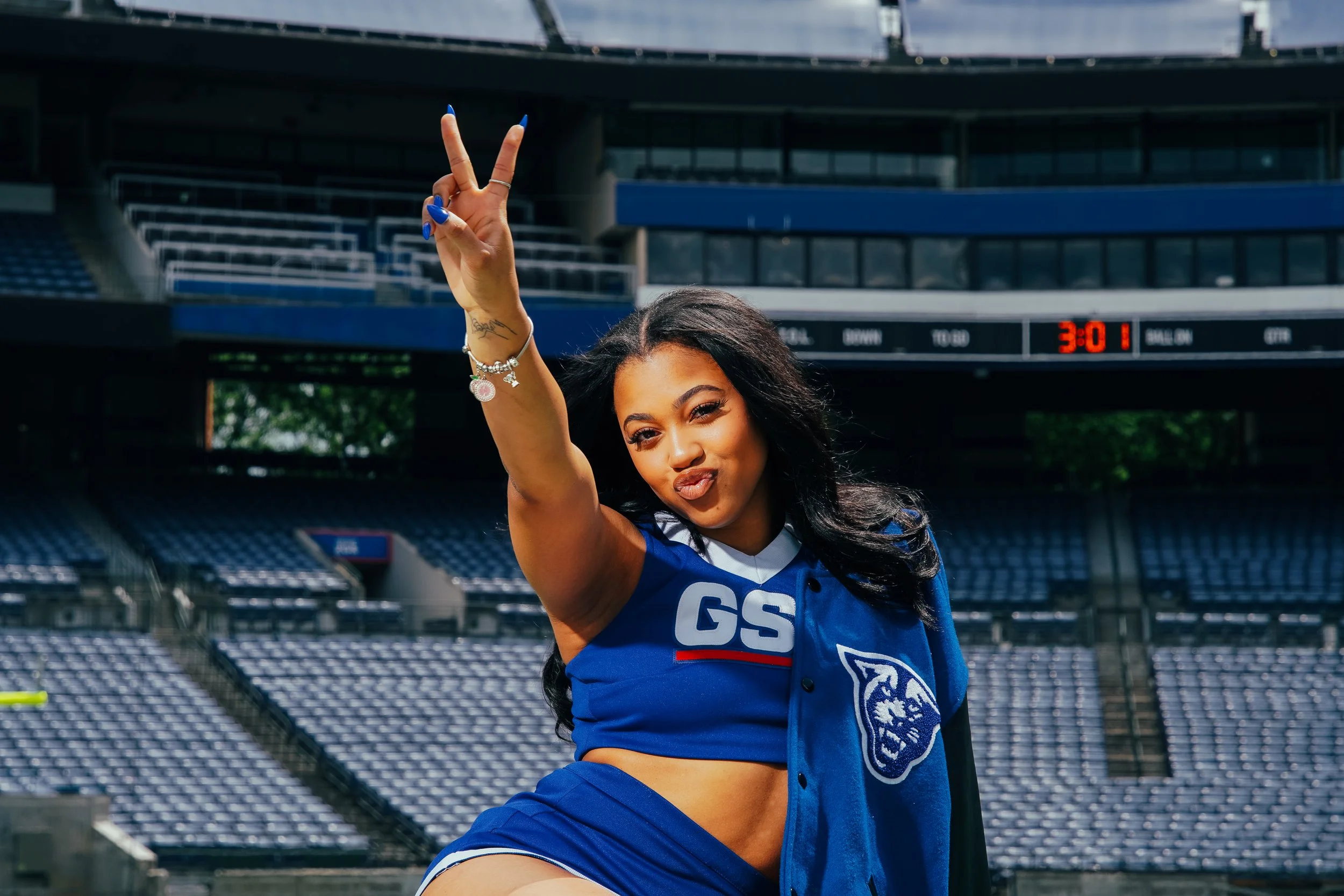 Woman in a blue sports uniform at a stadium, making a peace sign with her right hand, posing for the camera.