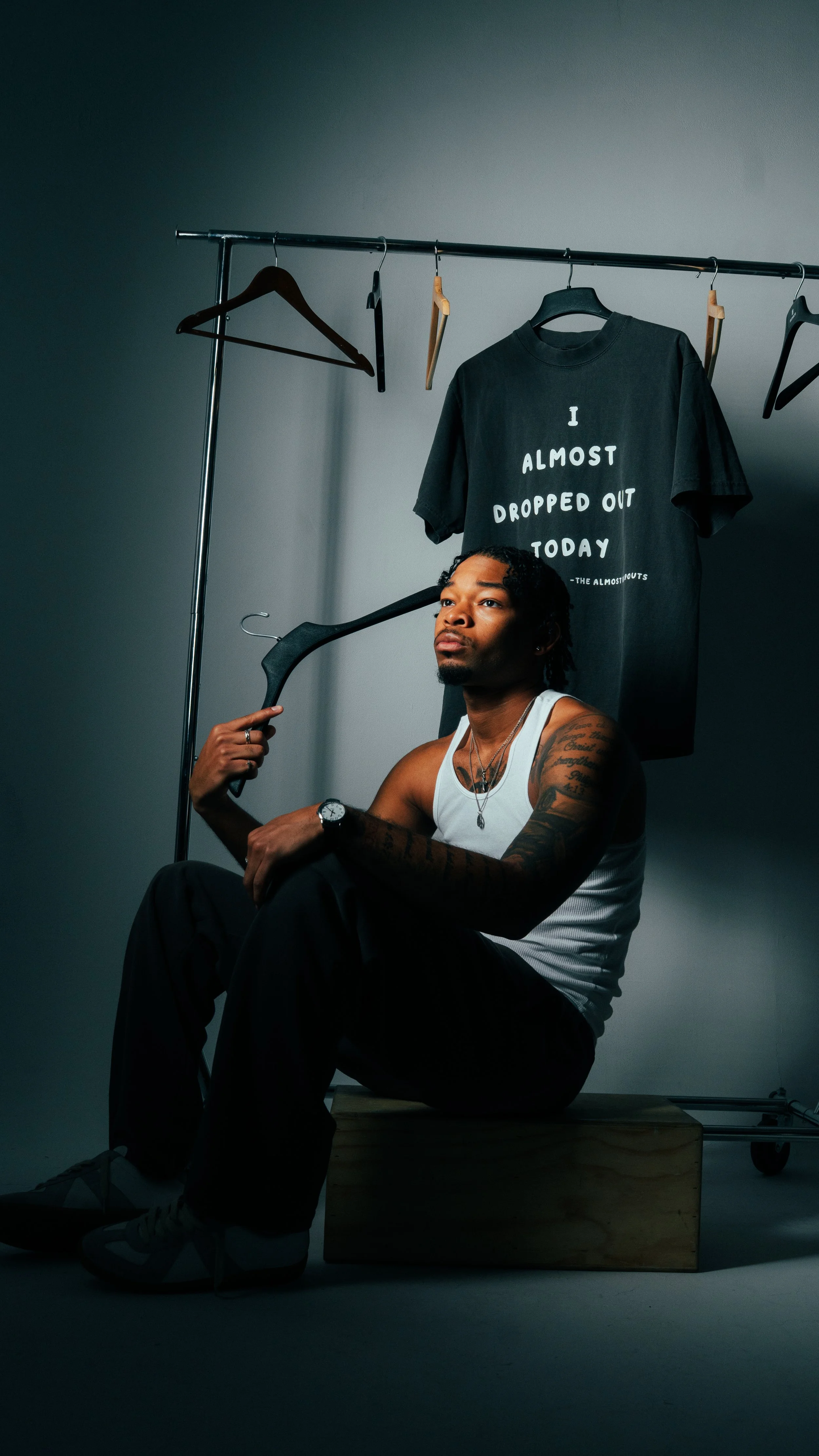 A man with tattoos wearing a white tank top, sitting on a wooden box, holding a hanger near his head, with a clothing rack and a black t-shirt that reads 'I ALMOST DROPPED OUT TODAY' hanging behind him.