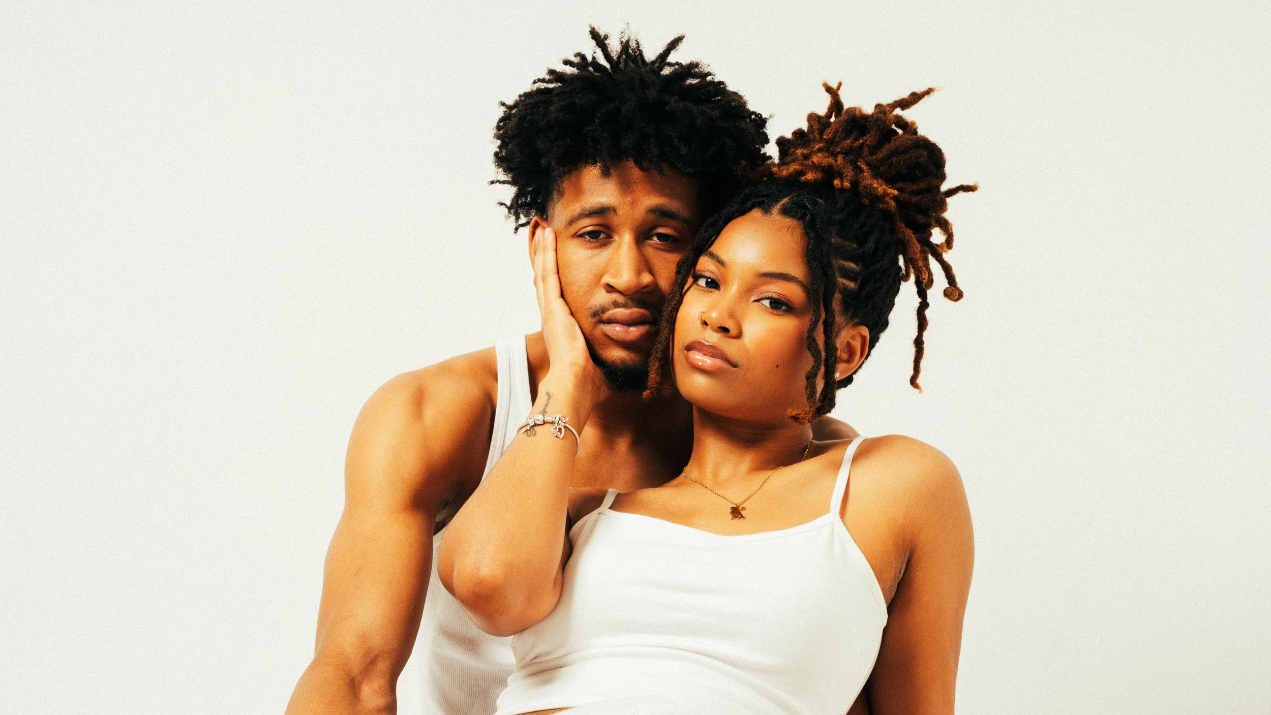 A young man and woman with dreadlocks posing closely together against a plain white background.