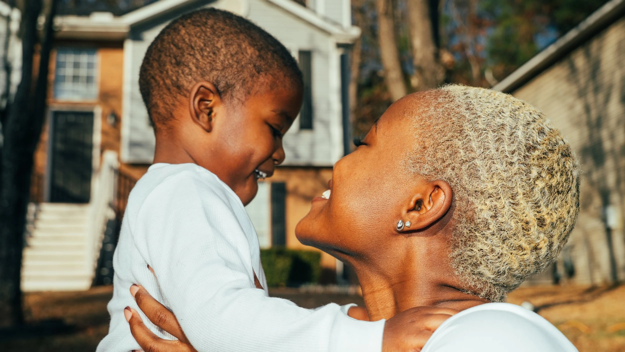 A young child and an older woman are embracing outdoors, smiling and looking at each other.