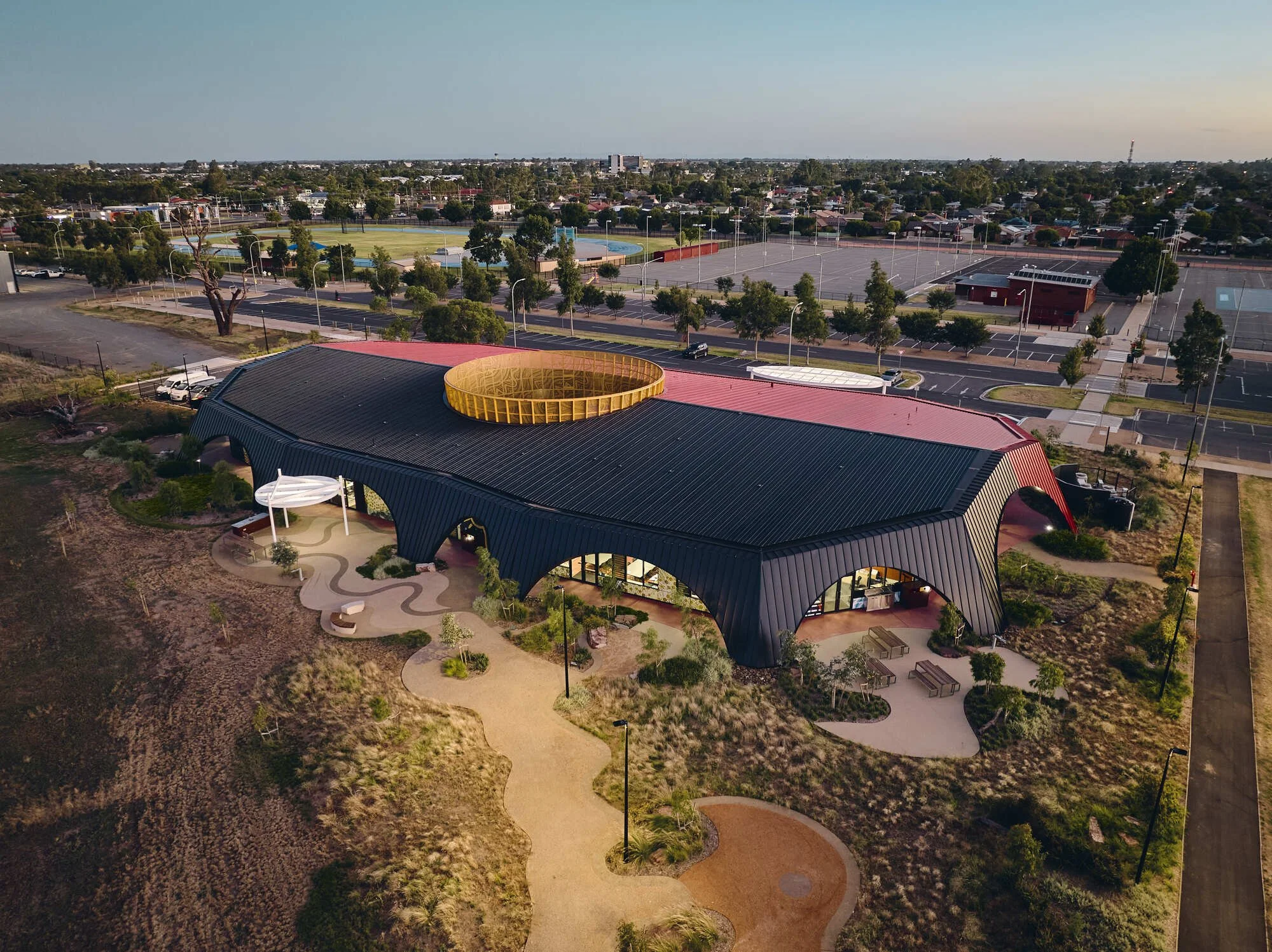 Aerial view of an indigenous modern building with black and red roof, circular opening on top, surrounded by a landscaped park with walking paths, benches, and greenery, in a suburban area with parking lots and sports fields in the background.