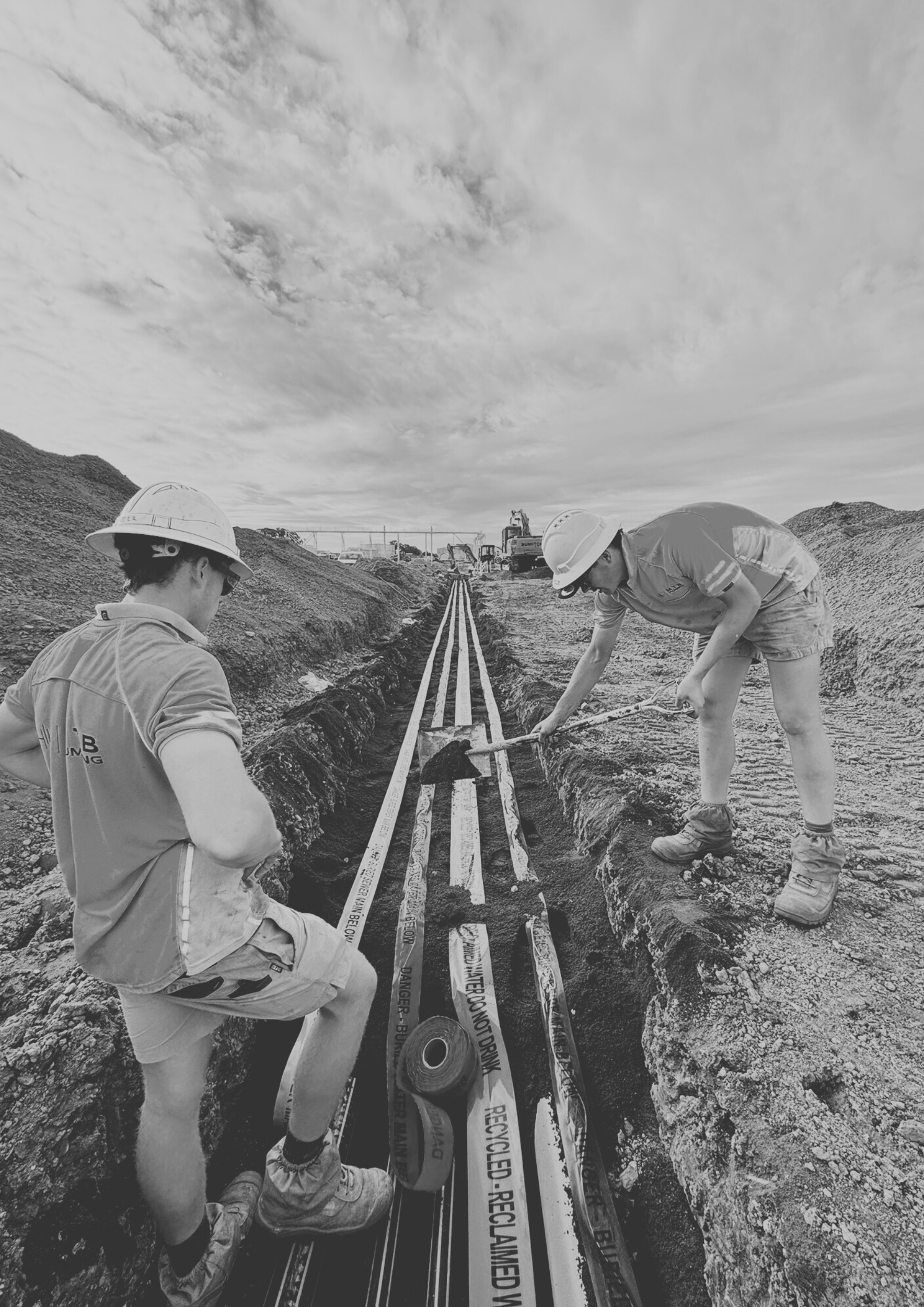 Two construction workers wearing hard hats working on laying underground cables or pipes in a trench at a construction site.