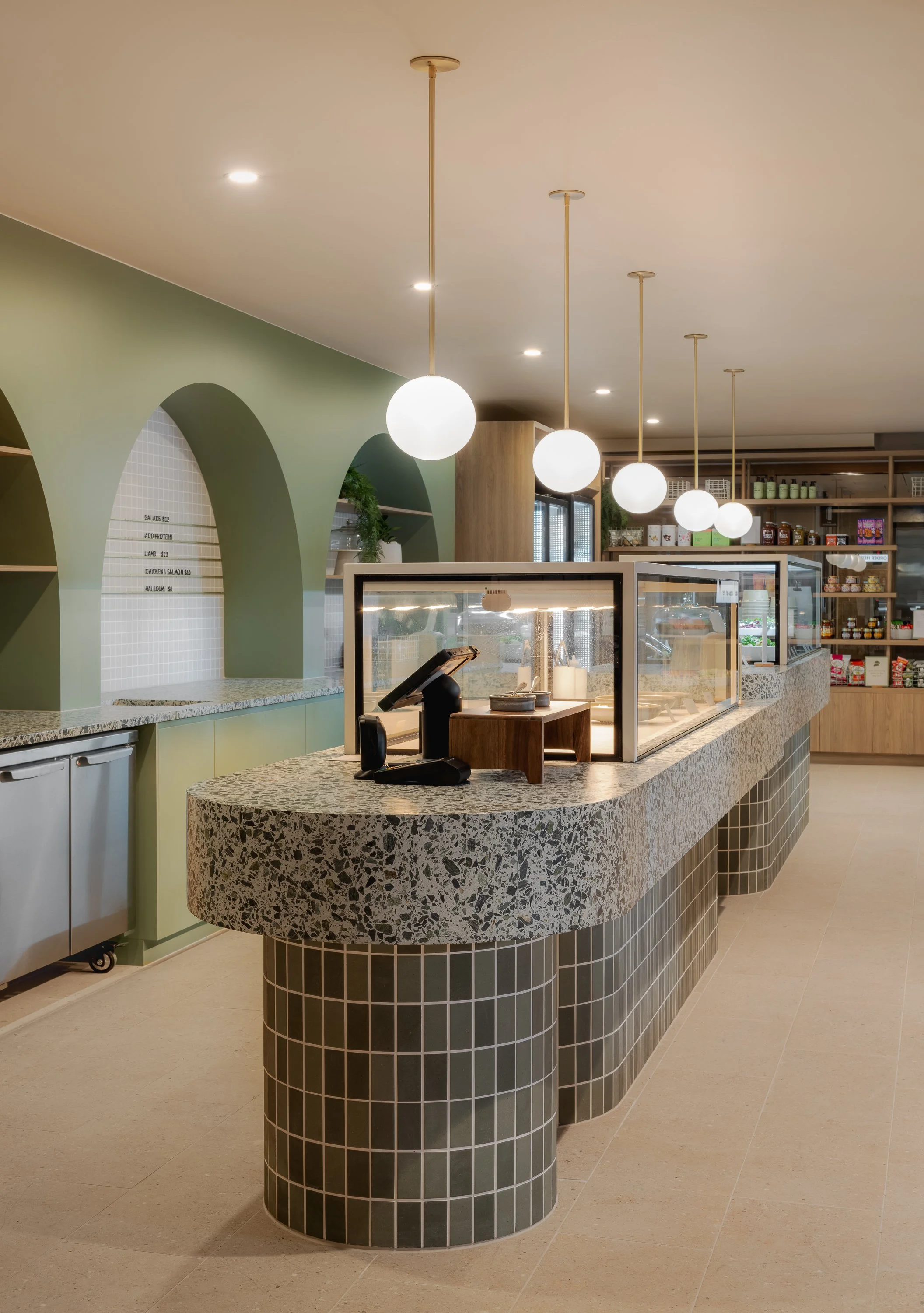 Curved cafe counter featuring green glazed tiles, terrazzo and suspended globe lighting.