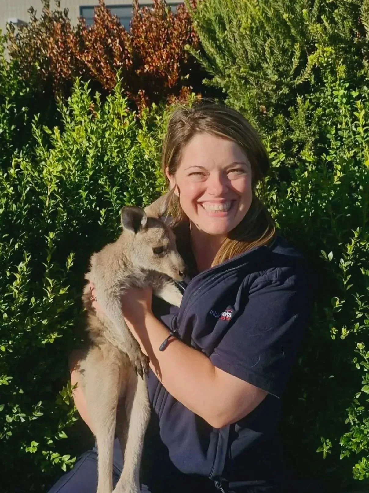 A woman smiling and holding a large cookie in her right hand. She is wearing a dark uniform jacket with a name badge, standing against a plain white wall.