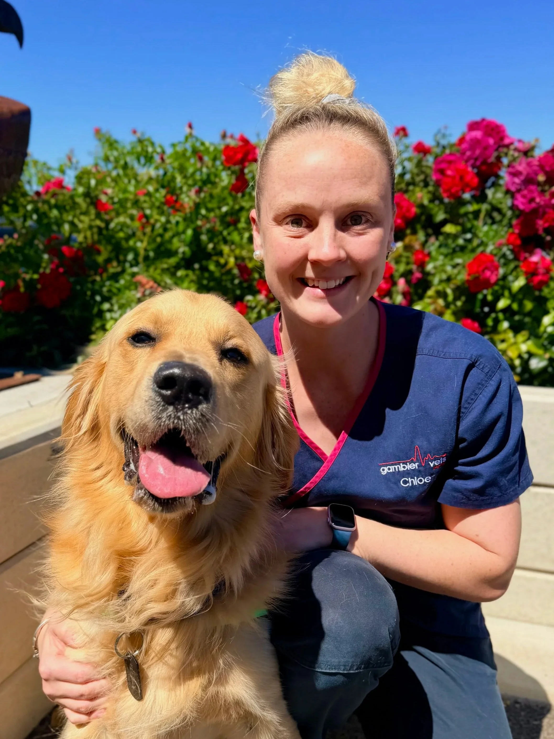 A smiling woman in navy scrubs standing outside of a vet clinic with a sign that reads 'Gambier Vets' and features a red heartbeat line graphic.