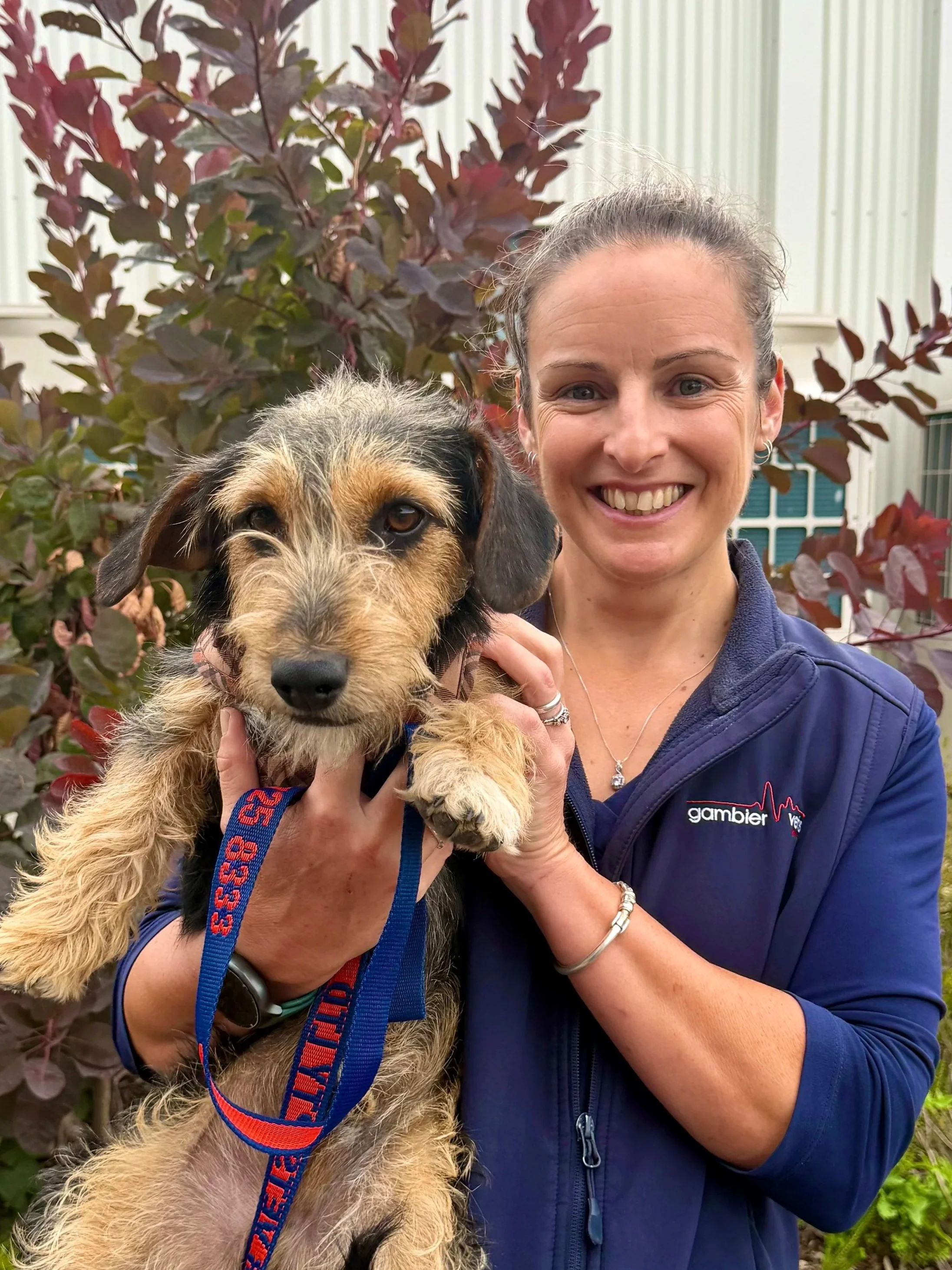 A woman smiling, wearing a black jacket with a Royal Canin logo, standing against a plain wall.
