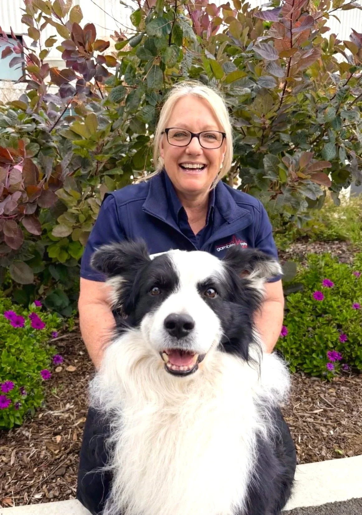 A woman with glasses and blonde hair smiling, wearing a navy blue uniform with a name tag and a logo, standing in front of a plant and a beige textured background.