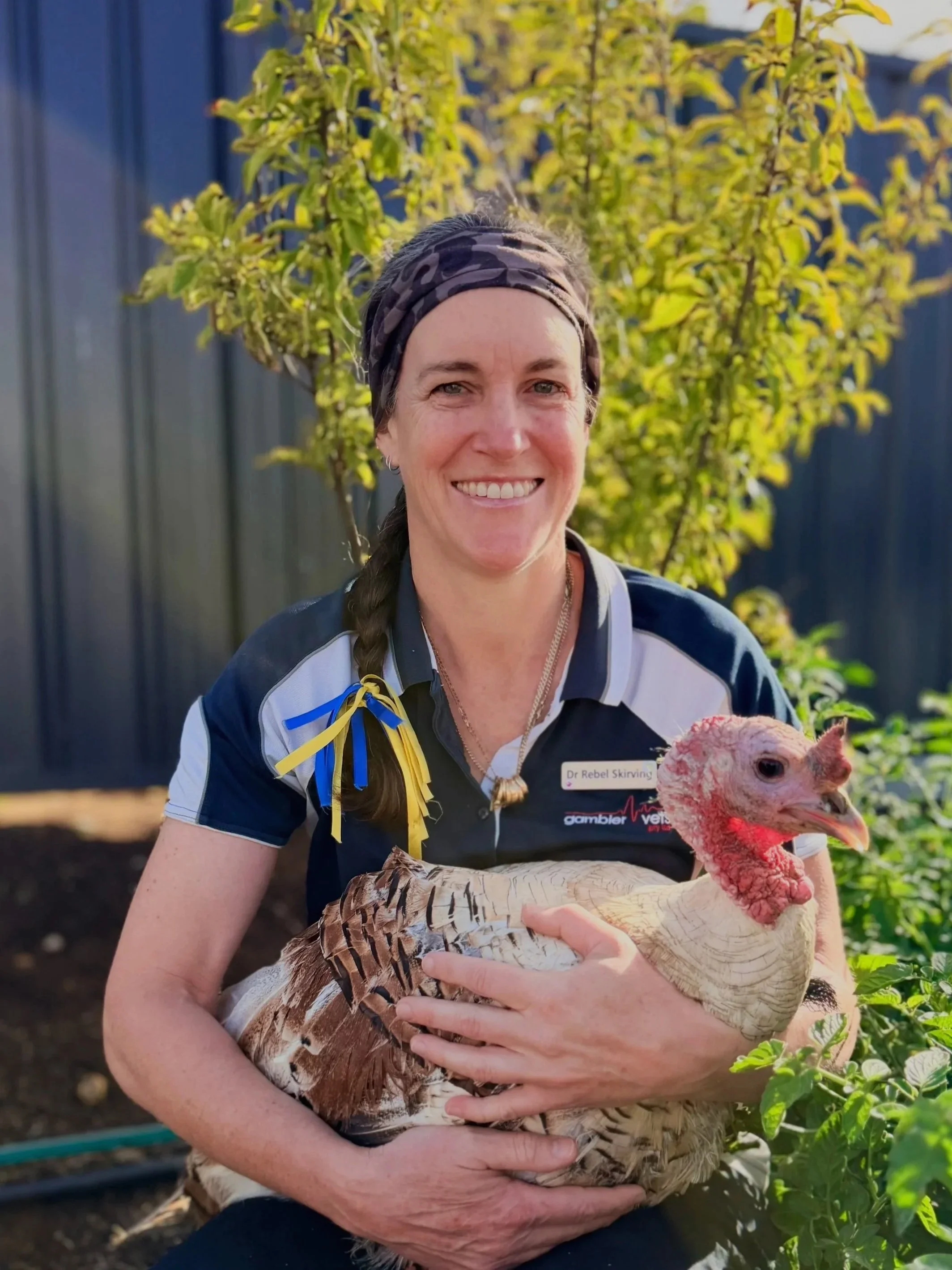 A young woman smiling outdoors, holding a chicken, with a dog nearby also looking at the chicken.