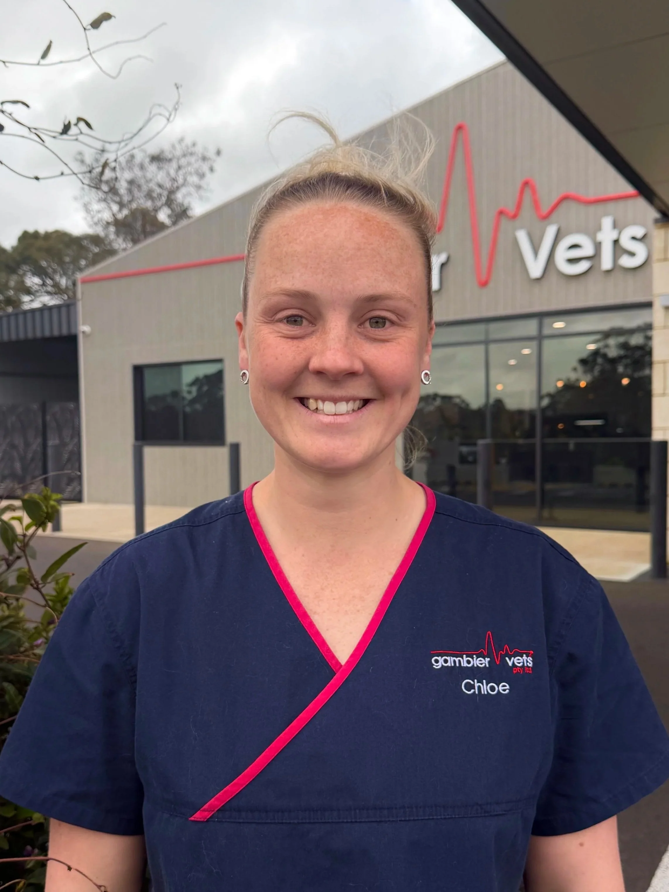 A smiling woman in navy scrubs standing outside of a vet clinic with a sign that reads 'Gambier Vets' and features a red heartbeat line graphic.