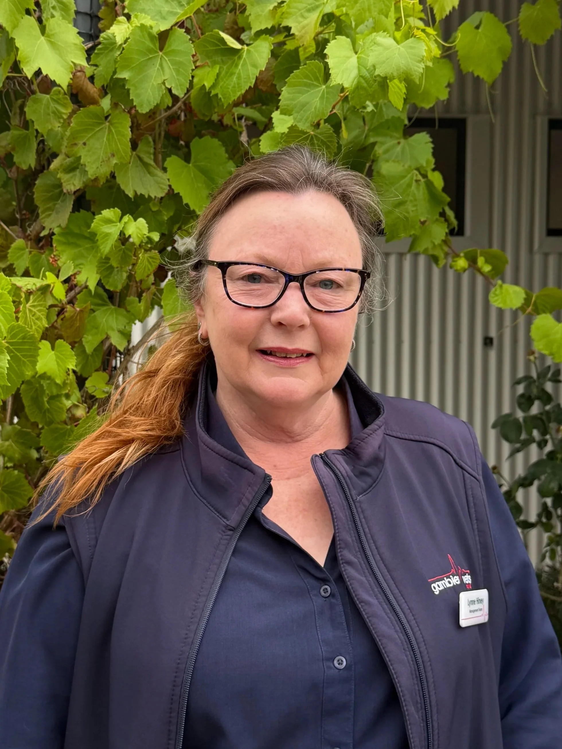 A smiling woman in a navy blue sweater with a name tag, standing against a plain light background.
