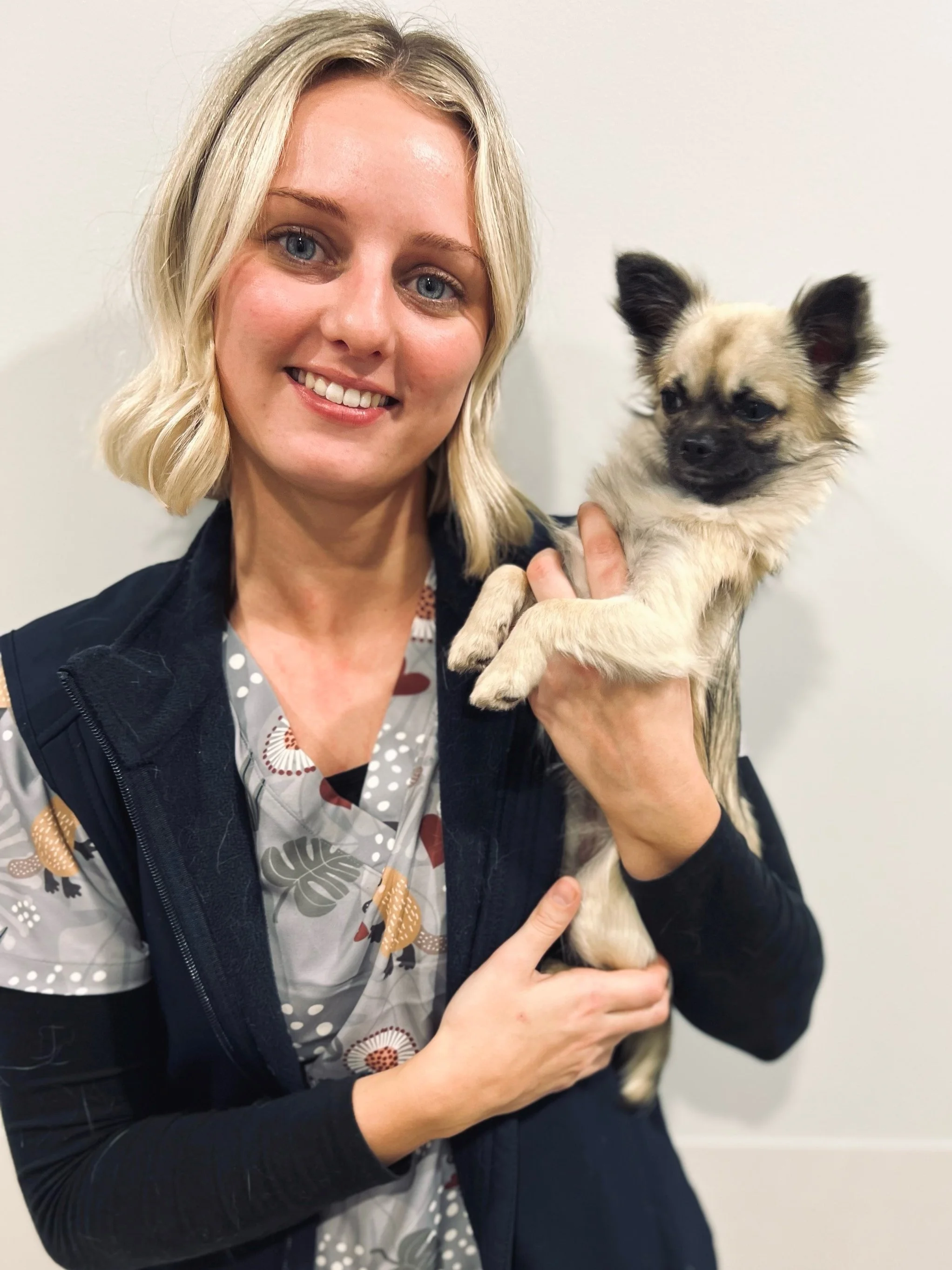 A woman with blonde hair smiling and holding a small fluffy dog with black facial markings, against a plain white wall.
