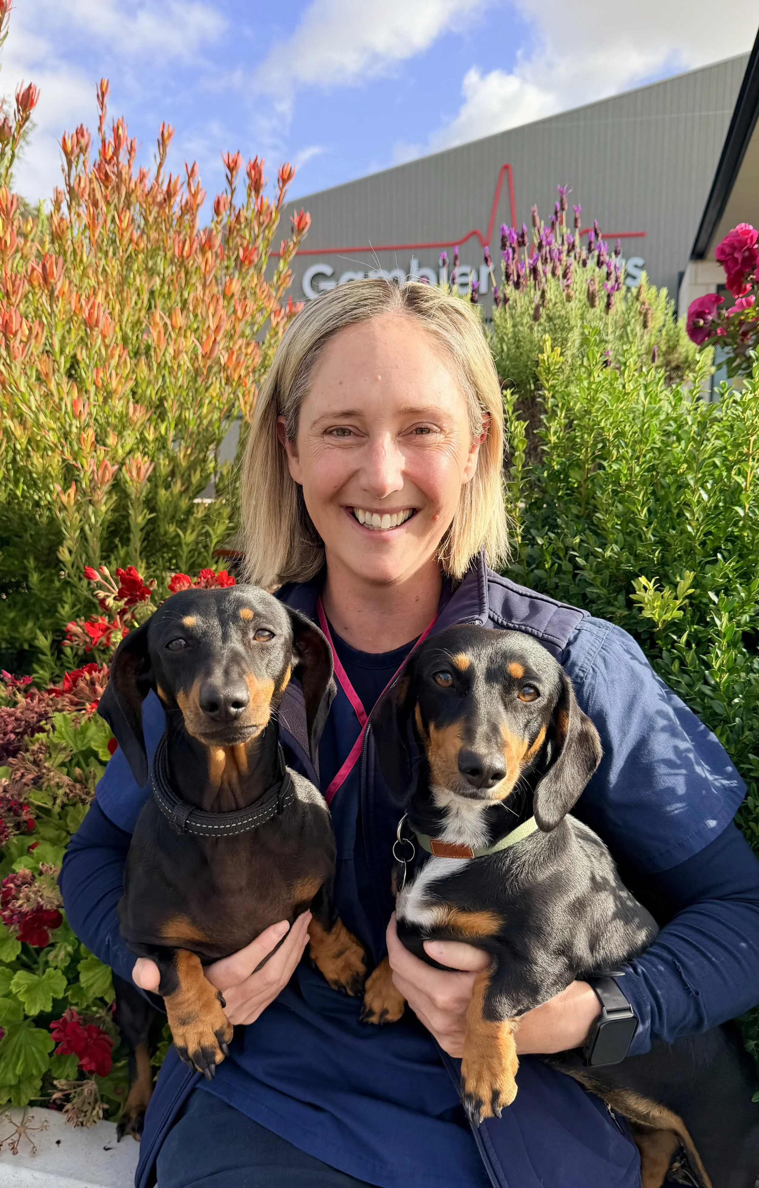 A woman with gray hair smiling, wearing a dark jacket, standing indoors near a window with plants outside.
