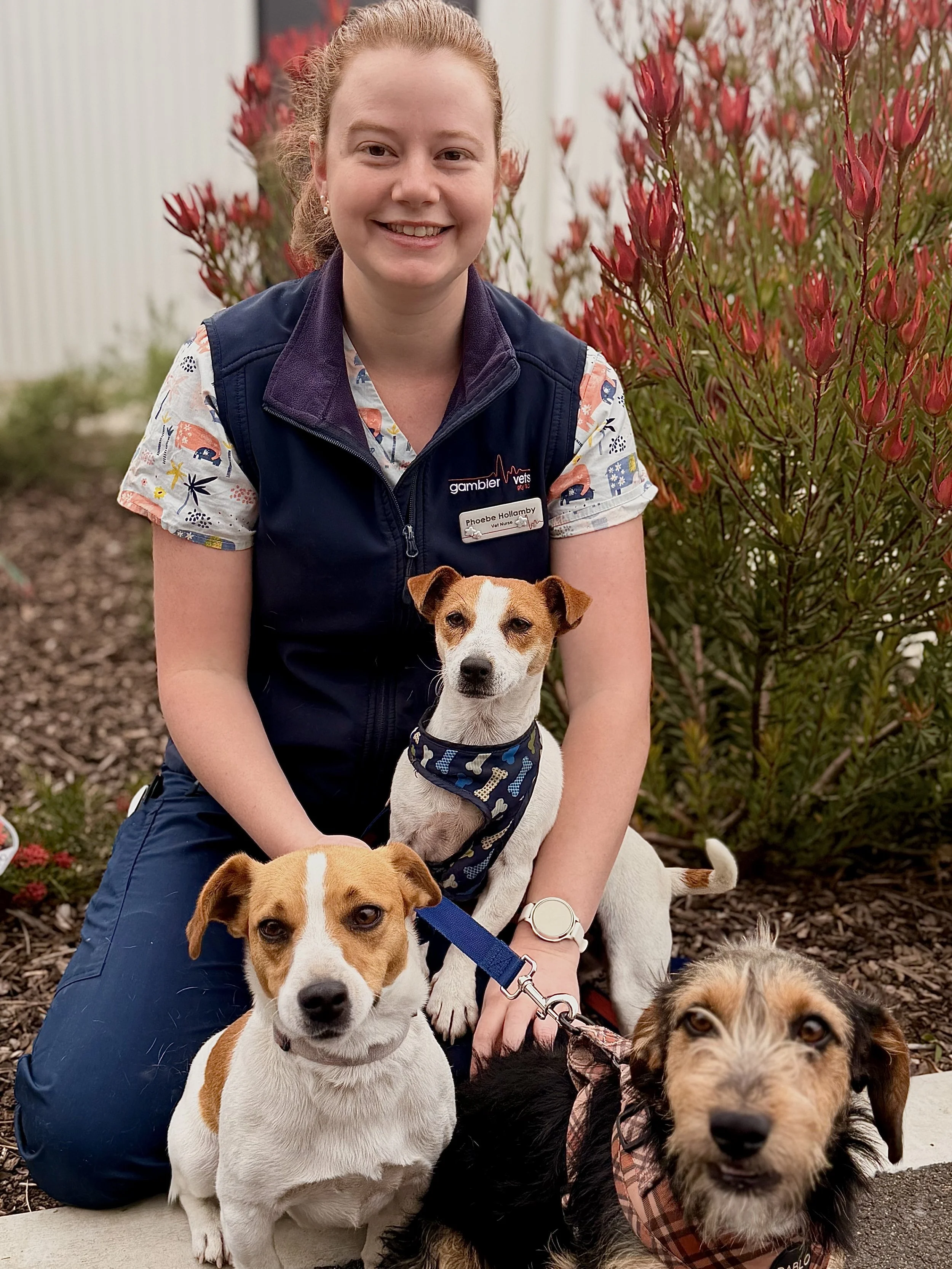 Young girl with red hair smiling, wearing a navy blue vest over a white shirt, standing against a plain off-white wall.