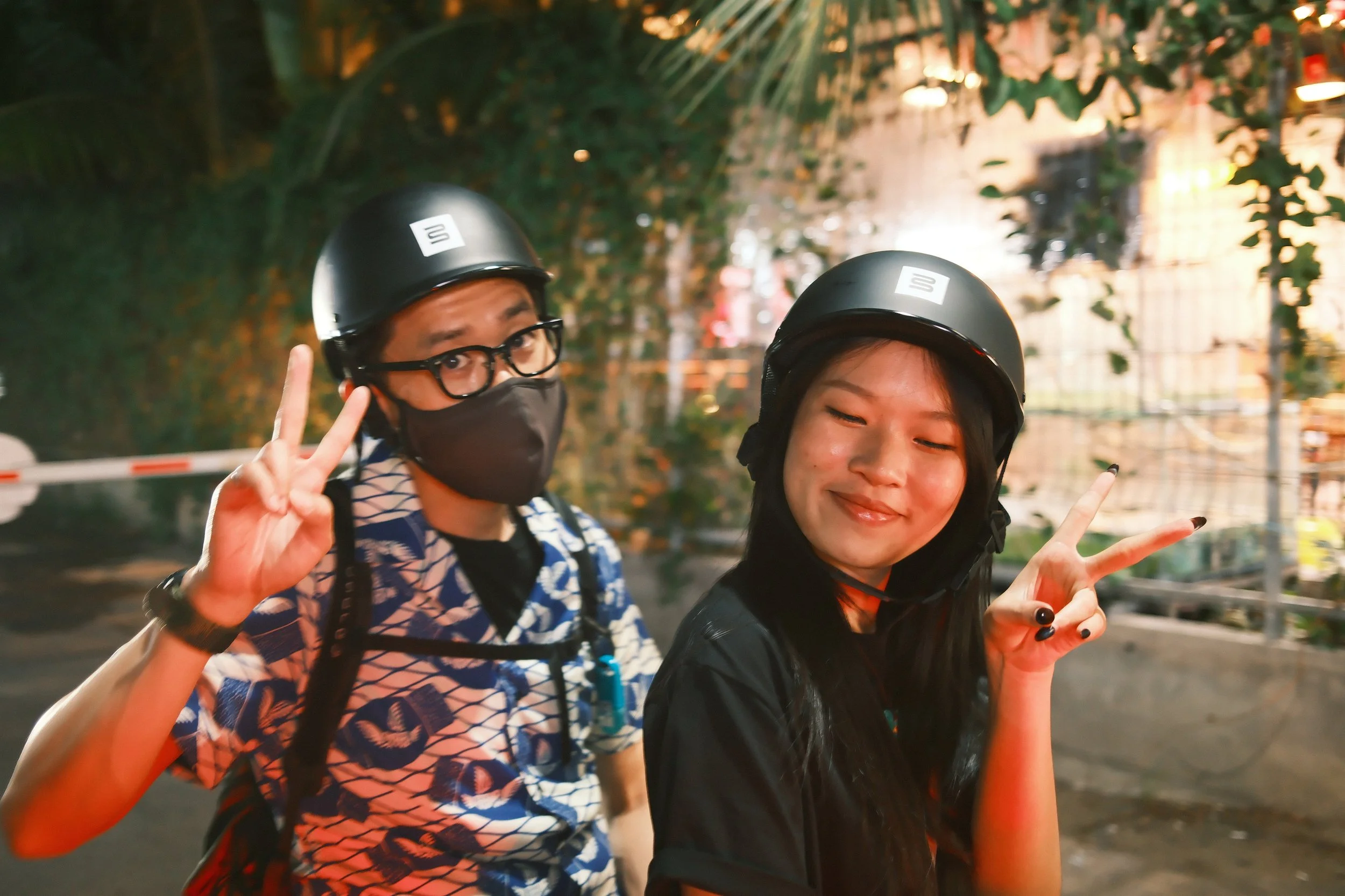 Two young people wearing black helmets, one with glasses and a face mask and the other with long black hair and closed eyes, smiling and making peace signs with their hands outdoors at night.