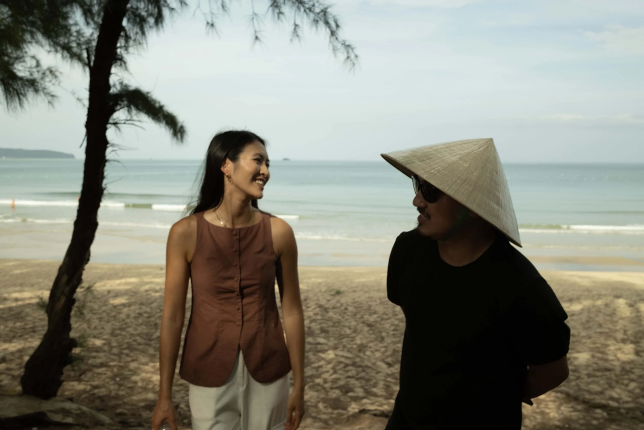 A woman and a man are standing on a beach, smiling and talking. The woman has long dark hair and is wearing a sleeveless brown top and white pants. The man is wearing sunglasses, a traditional Asian conical hat, and a black shirt. There are trees and