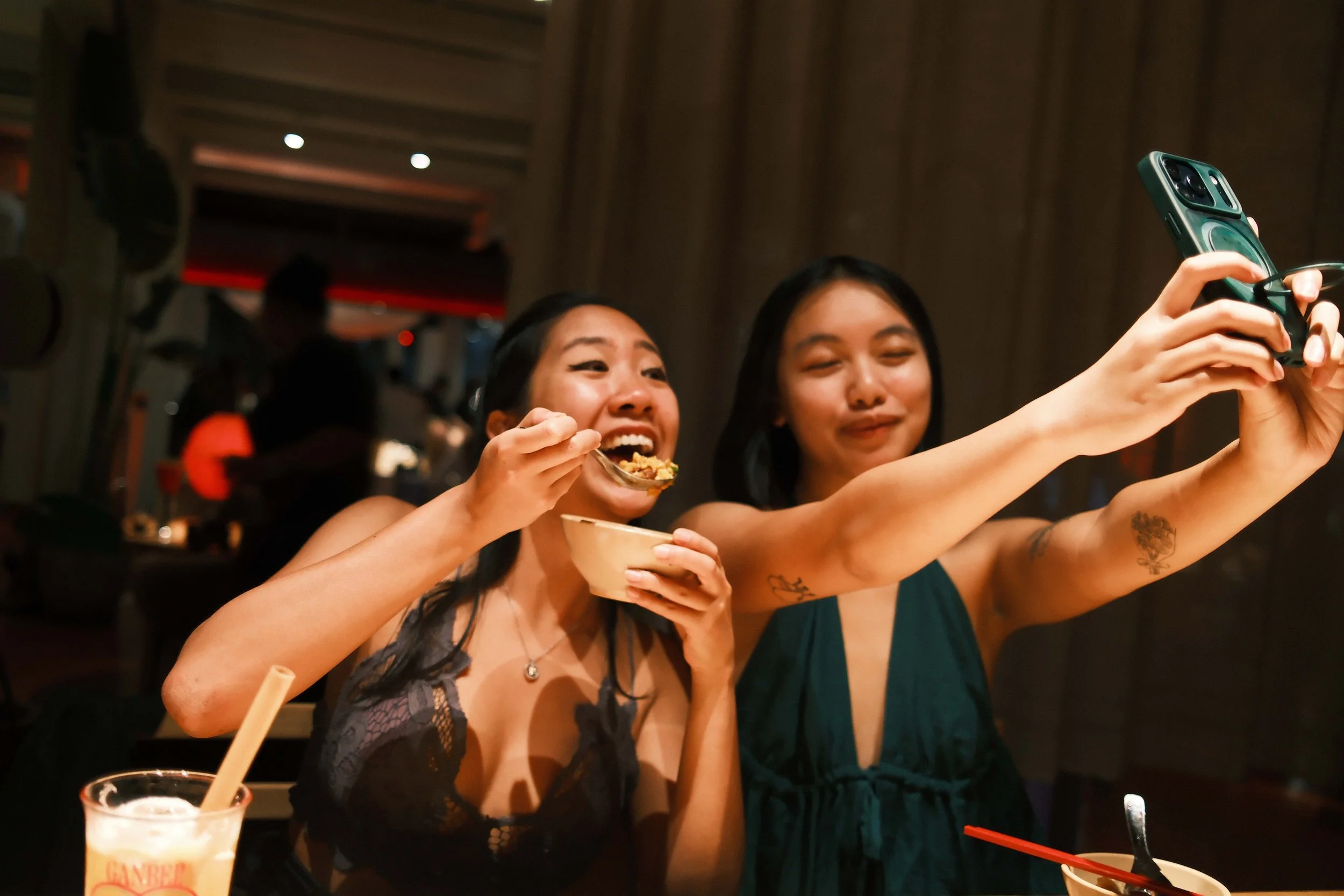 Two women taking a selfie while dining at a restaurant. One woman is eating food from a bowl, and the other is holding a smartphone to capture the photo.