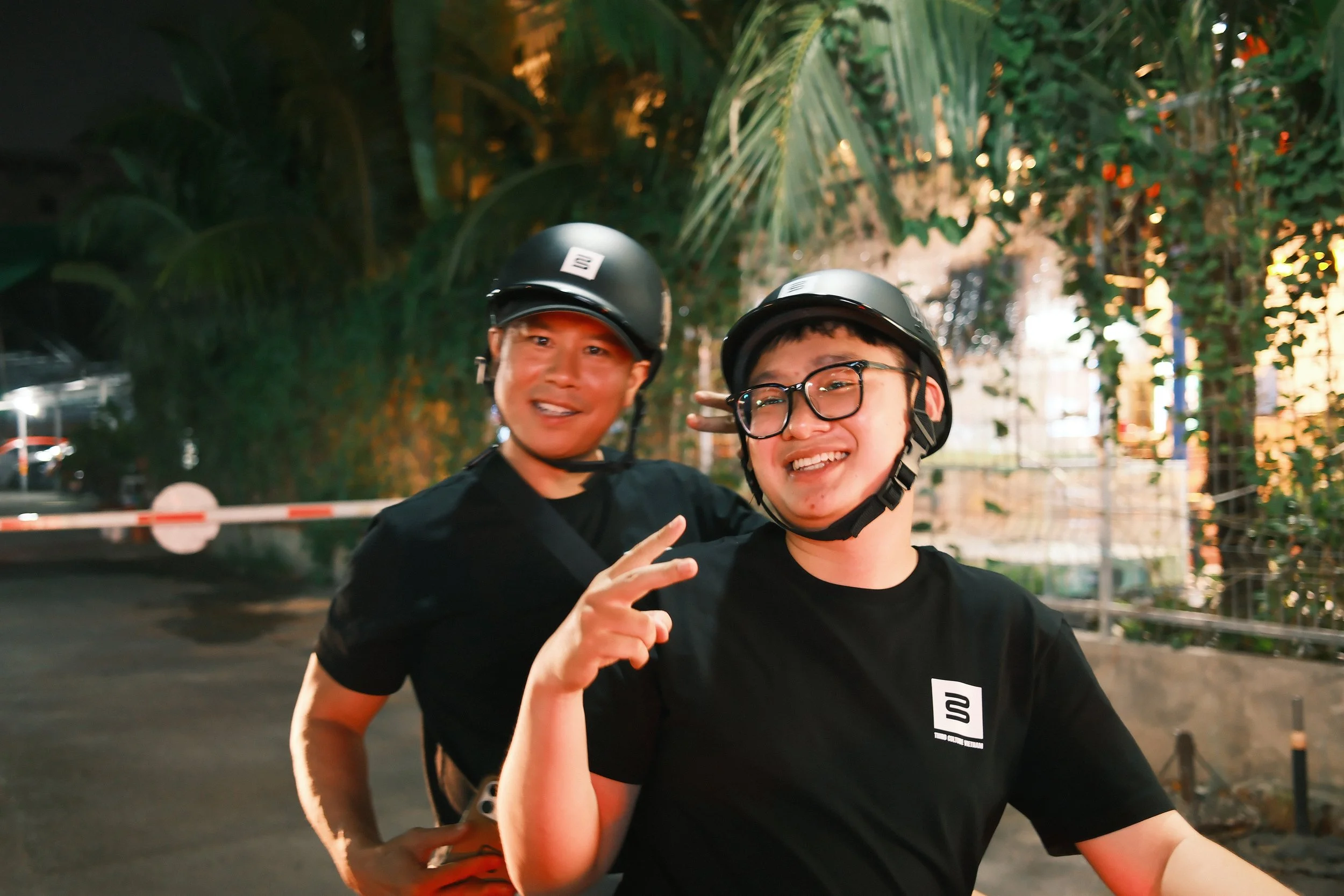Two young men wearing black t-shirts and helmets, smiling for a photo outdoors in evening with trees and lights in the background.