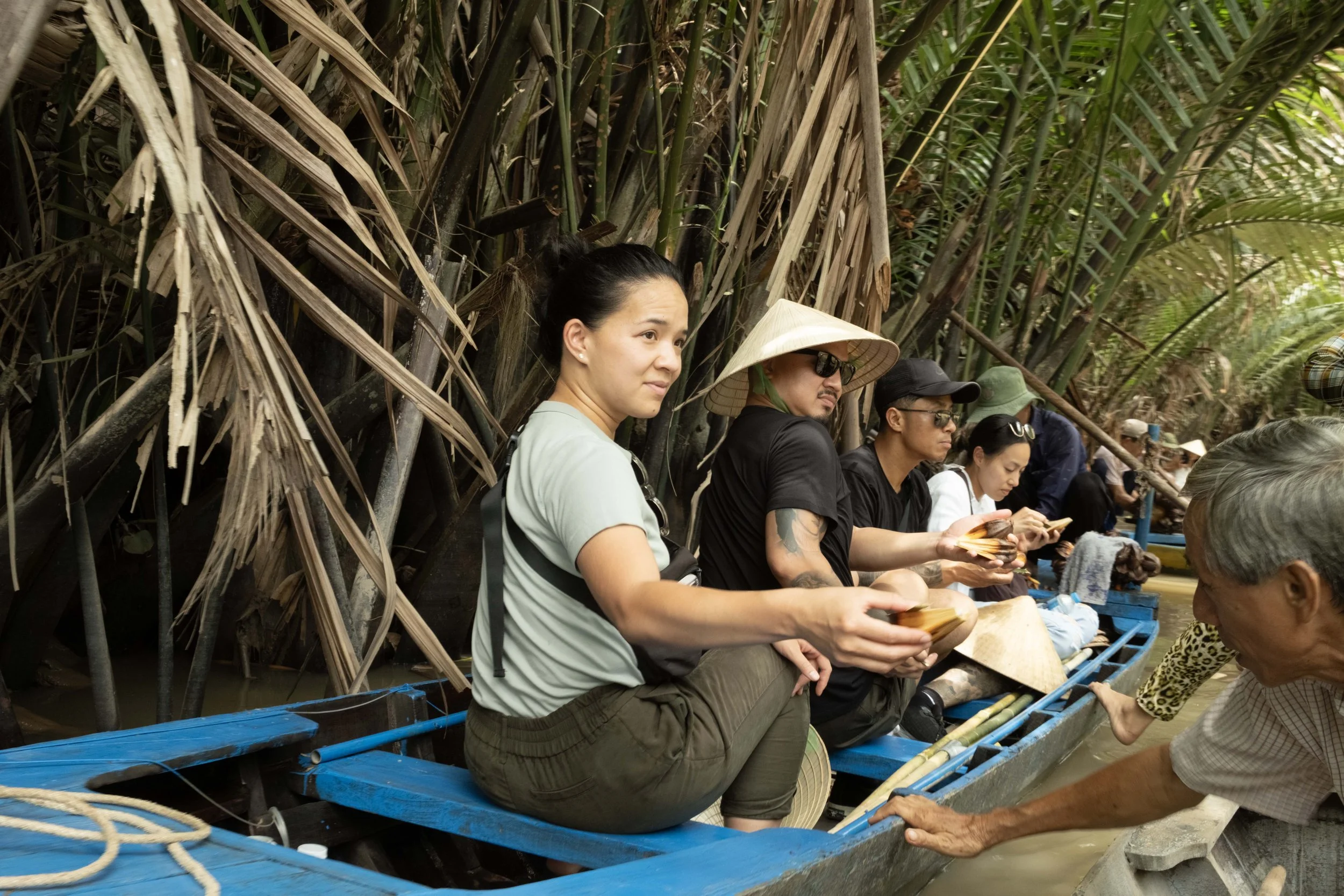 Tourists sitting in a boat on a river surrounded by dense tropical foliage, some wearing traditional conical hats, while an elderly local man leans into the boat.