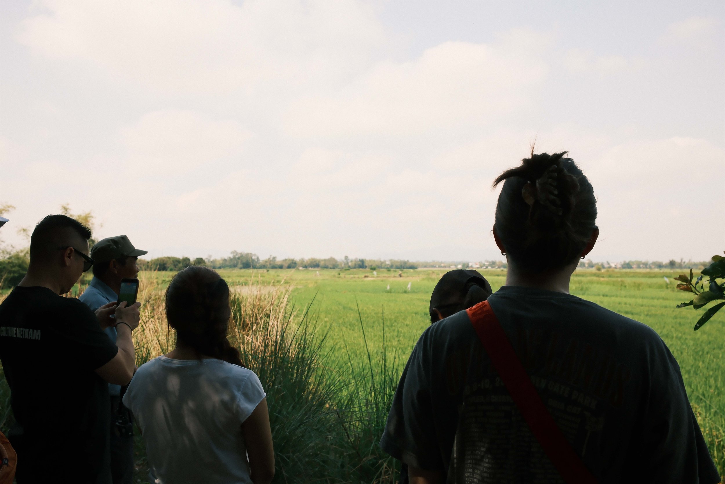 Group of people standing outdoors in front of green fields, facing away from the camera, with a cloudy sky above.
