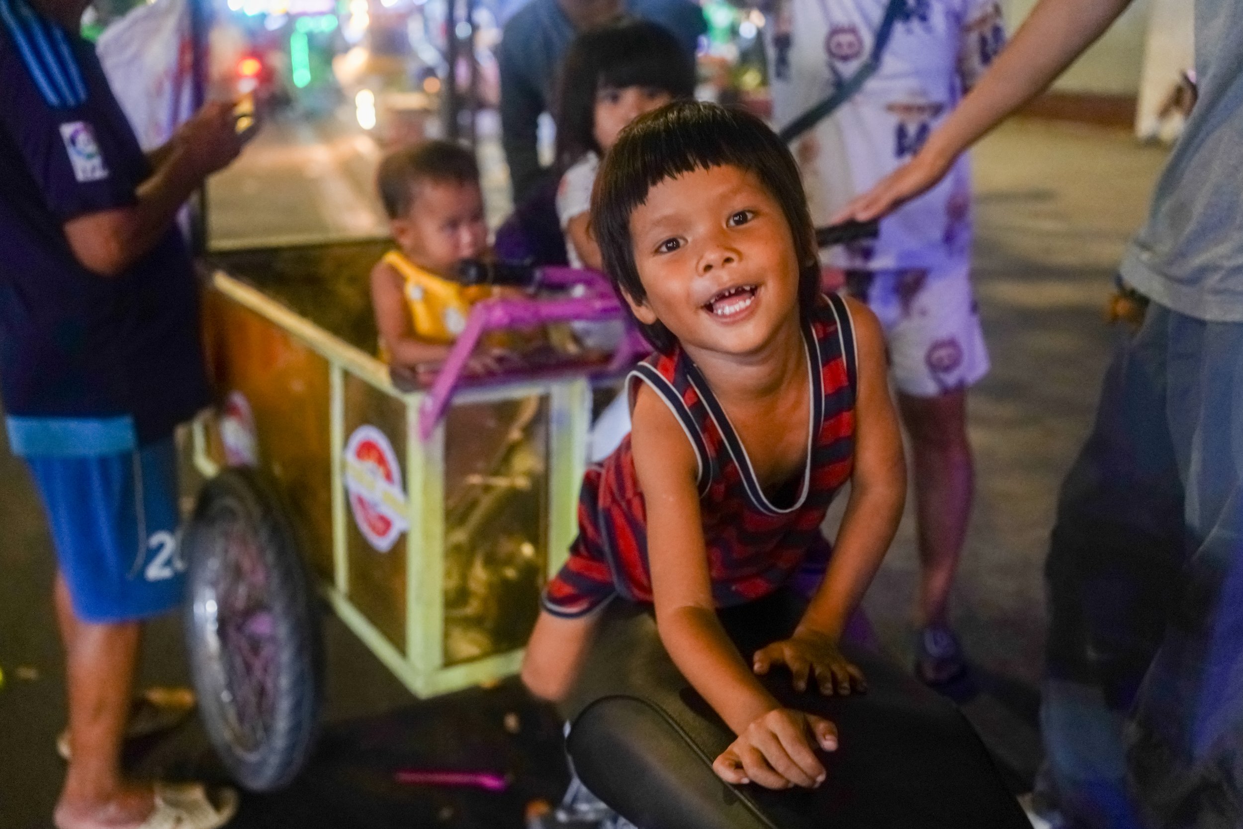A young boy with a grin, on hands and knees, is on a motorcycle in a busy street at night with children, adults, and street lights in the background.