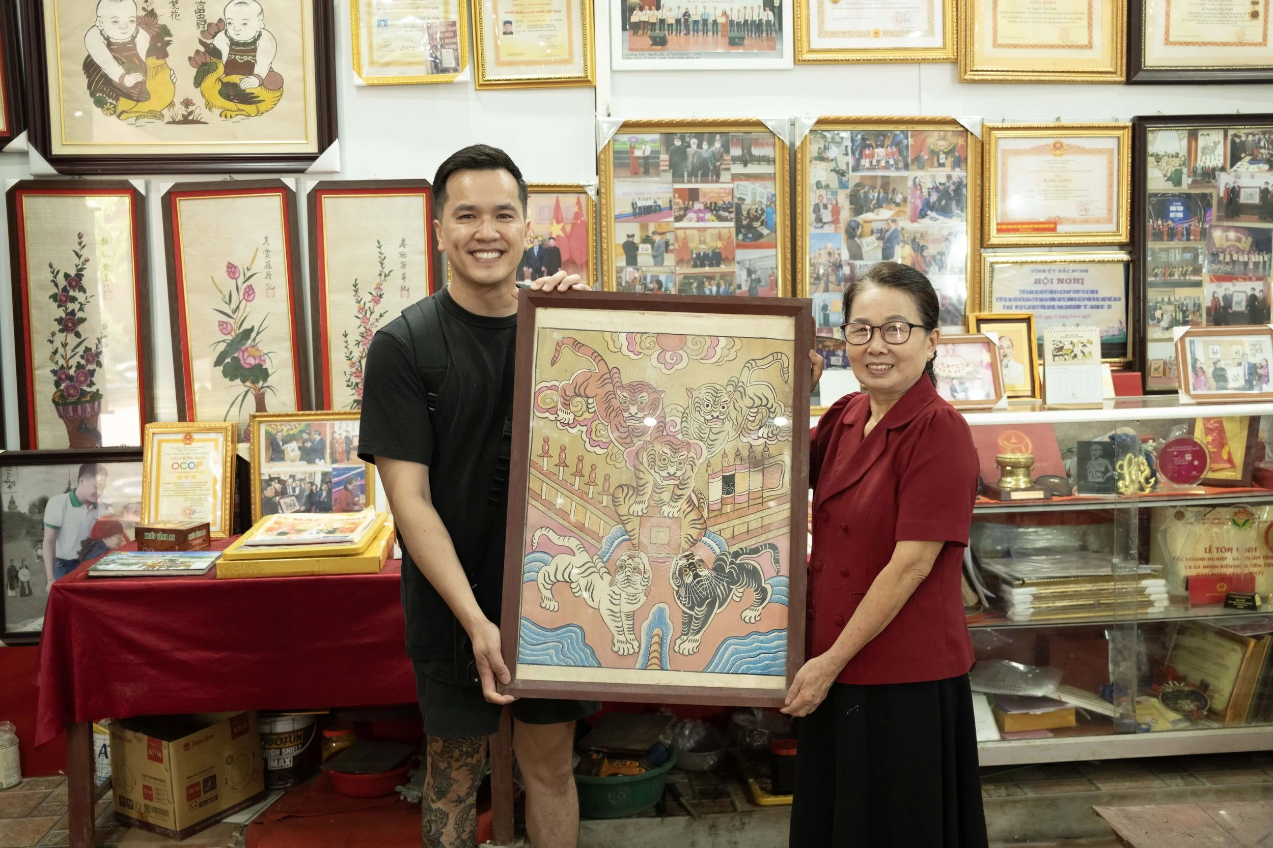 A man and a woman are smiling and holding a framed artwork of tigers and clouds. They are in a room with numerous framed photographs, certificates, and artwork on the walls and shelves behind them.