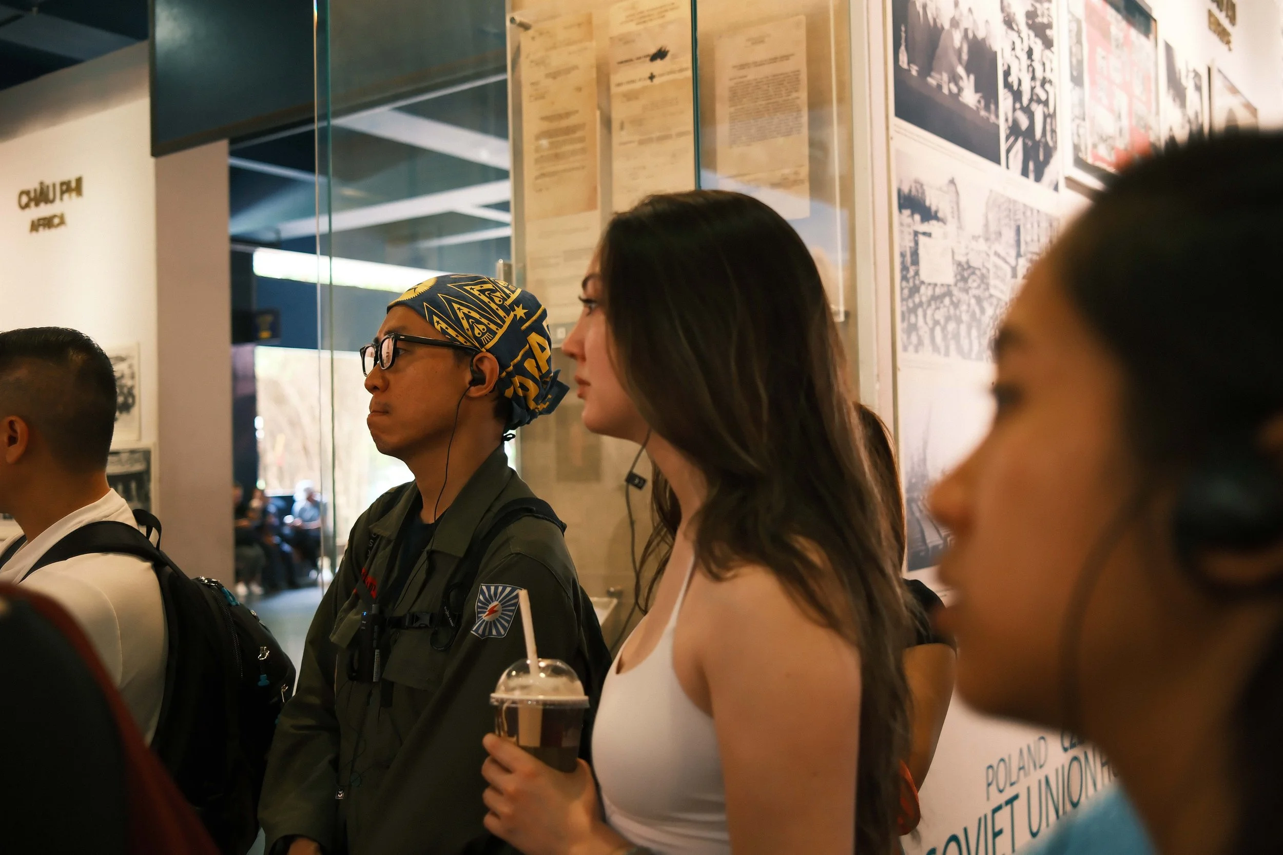 A diverse group of people standing in line at an indoor event or exhibition, with various posters and informational displays on the wall behind them. One woman is holding a drink in a plastic cup, and some individuals are wearing headphones or earbud