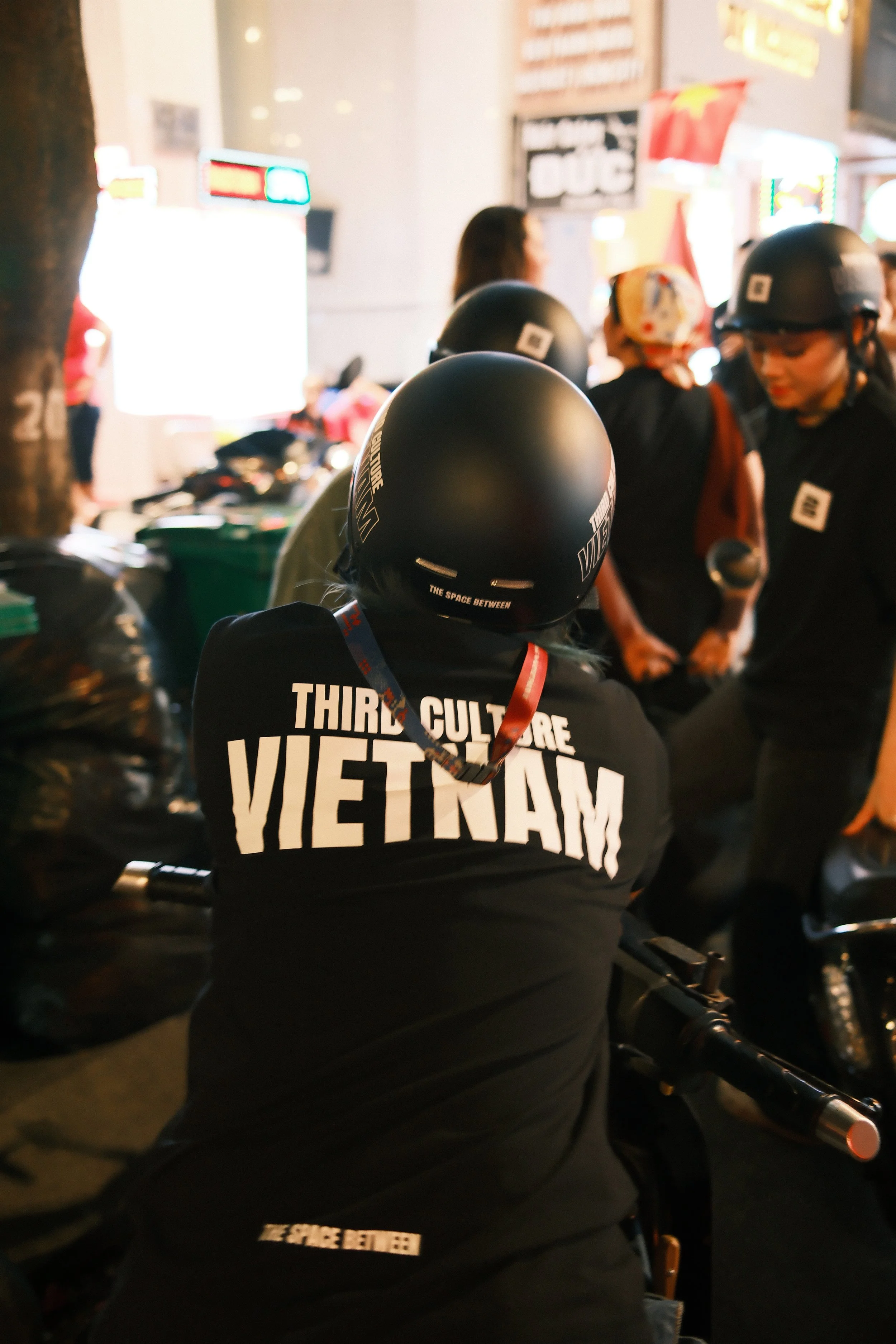 Person wearing a helmet and a black jacket with the words "Third Culture Vietnam" and "The Space Between" printed on the back, sitting on a motorcycle in a busy street at night, with other people and bright lights in the background.