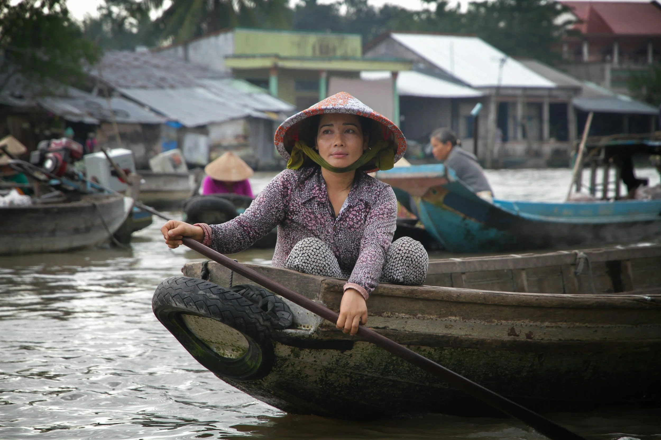 A woman with a traditional conical hat sitting in a small boat on a river, with other boats and houses in the background.