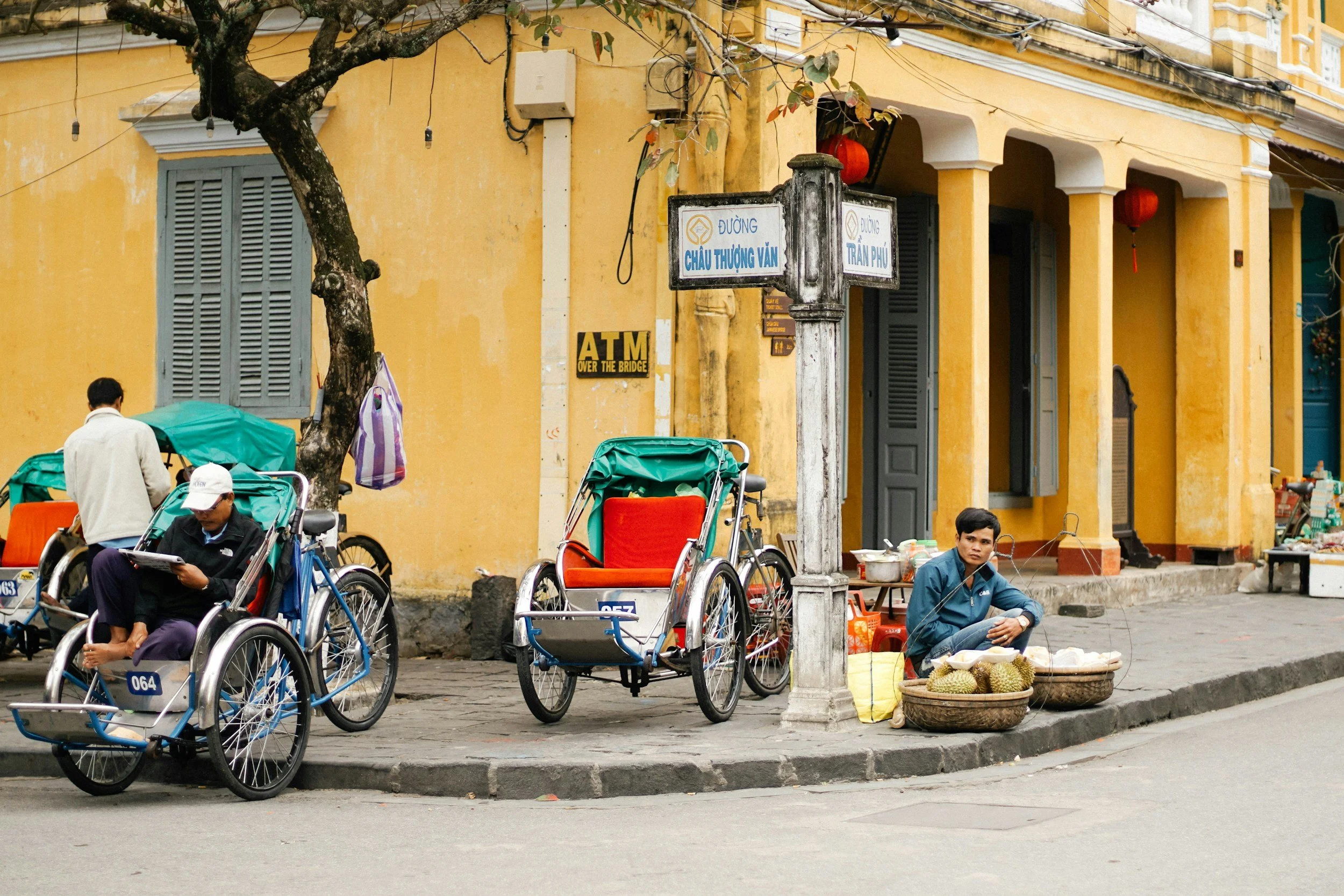 Street scene with two rickshaws parked on a sidewalk, a man sitting on the ground with baskets of durian, and a person walking past. Yellow buildings and street signs are visible.