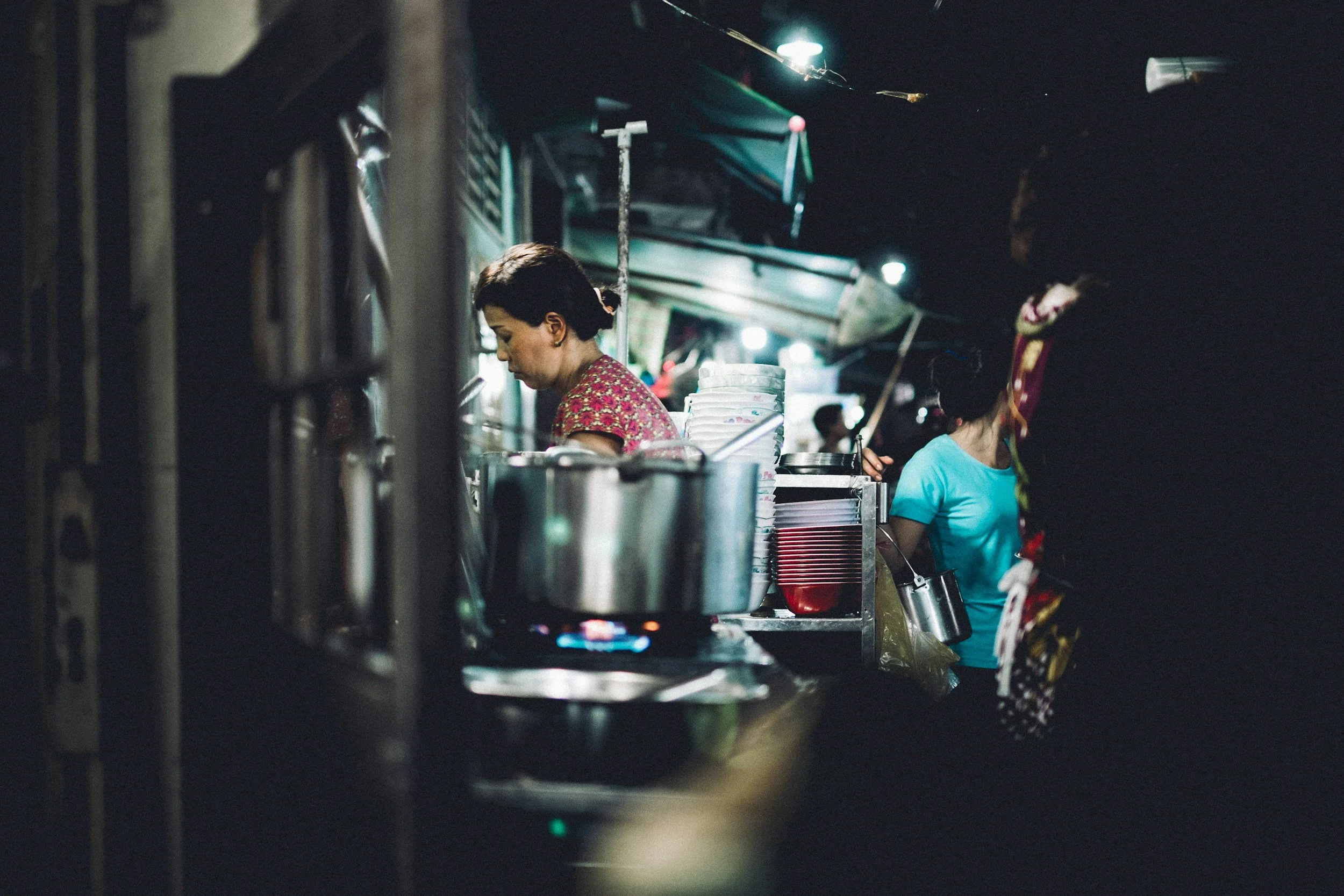 Two women working in a street food stall at night, with kitchen utensils, containers, and cooking equipment visible.