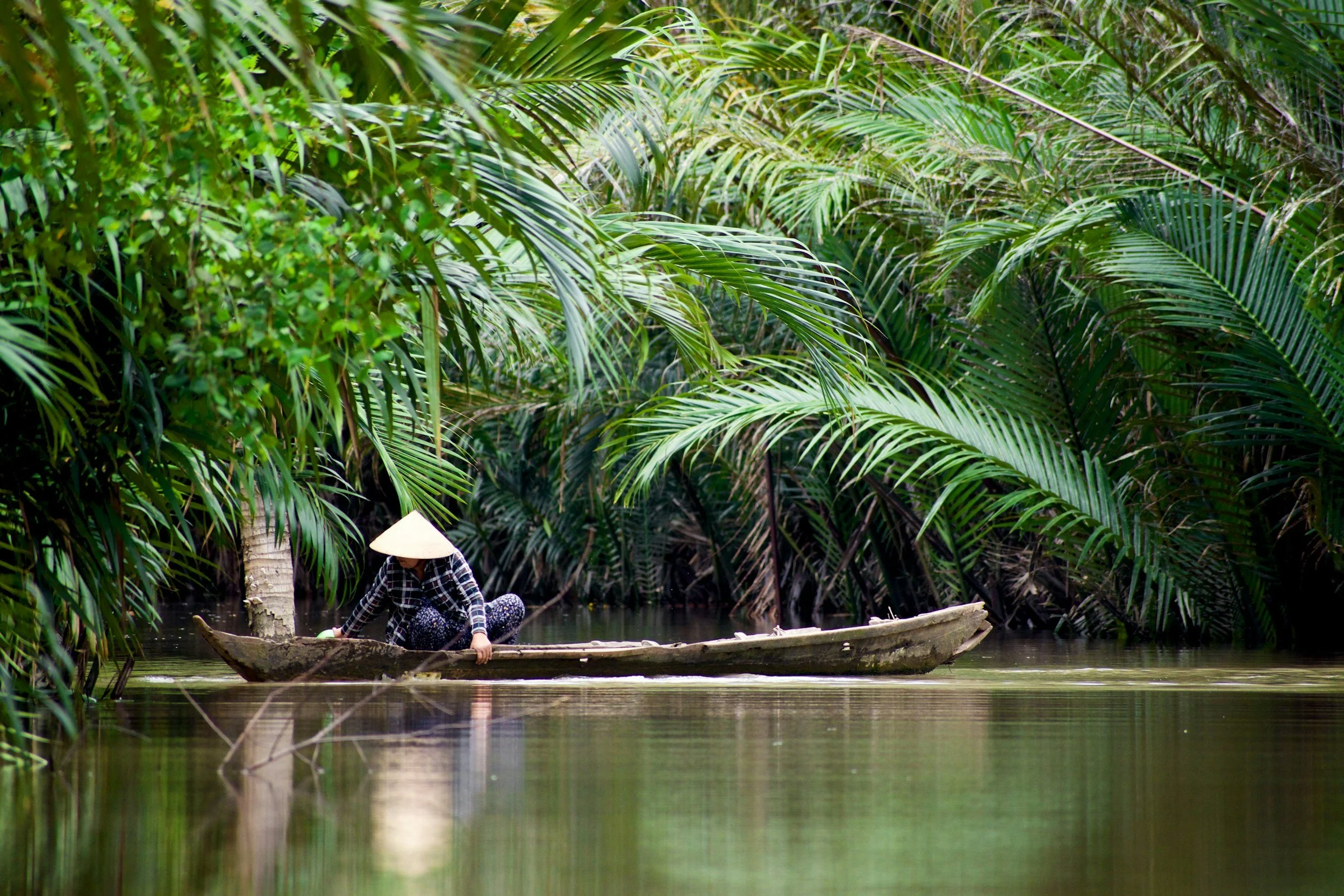A person wearing a traditional Asian conical hat and dark clothing is paddling a small wooden boat through a narrow, tranquil waterway surrounded by dense, lush green tropical foliage.