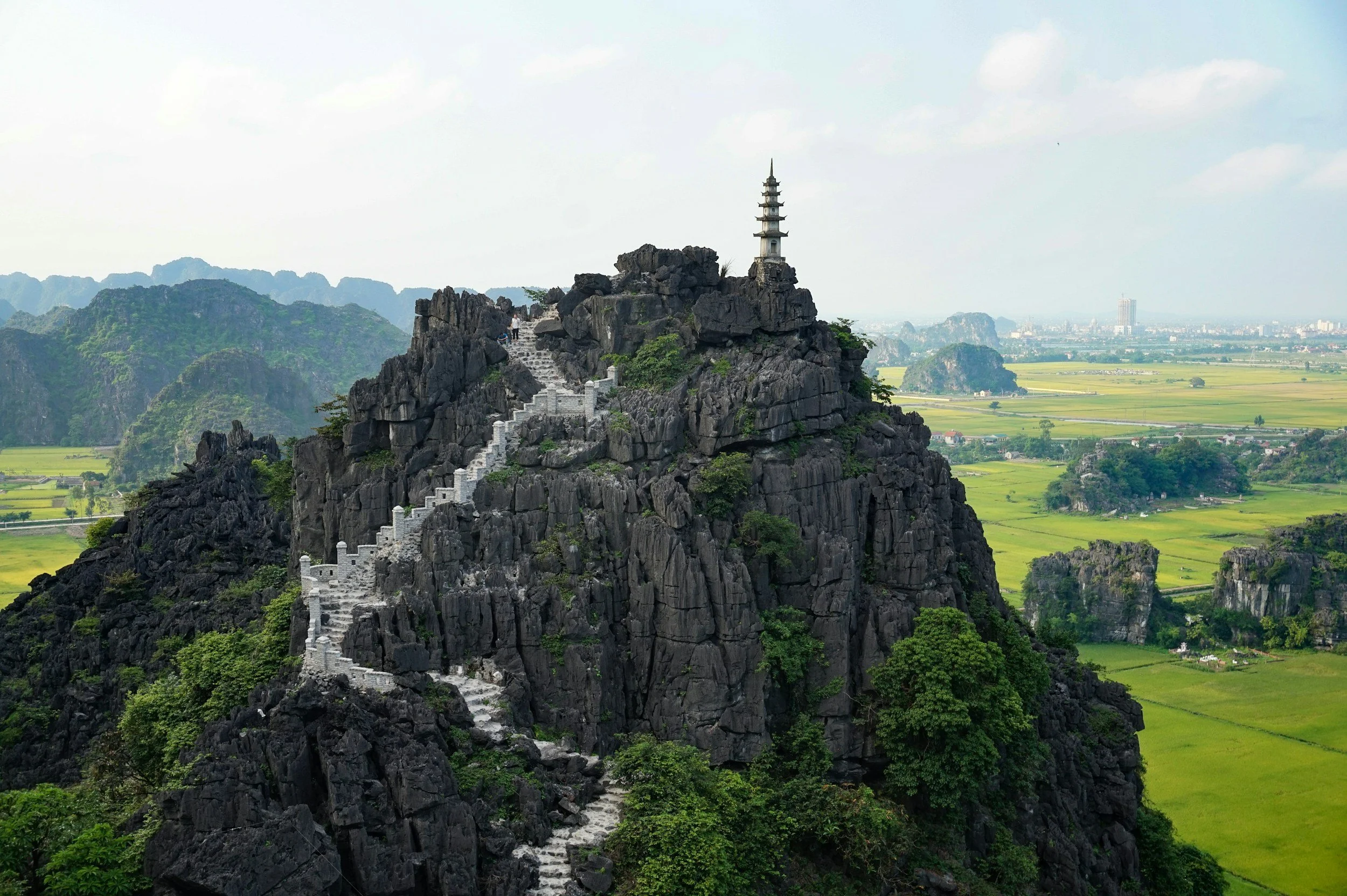 A rocky mountain with a pagoda on top, and a staircase winding up the mountain. The background shows a lush green landscape with fields and distant mountains.