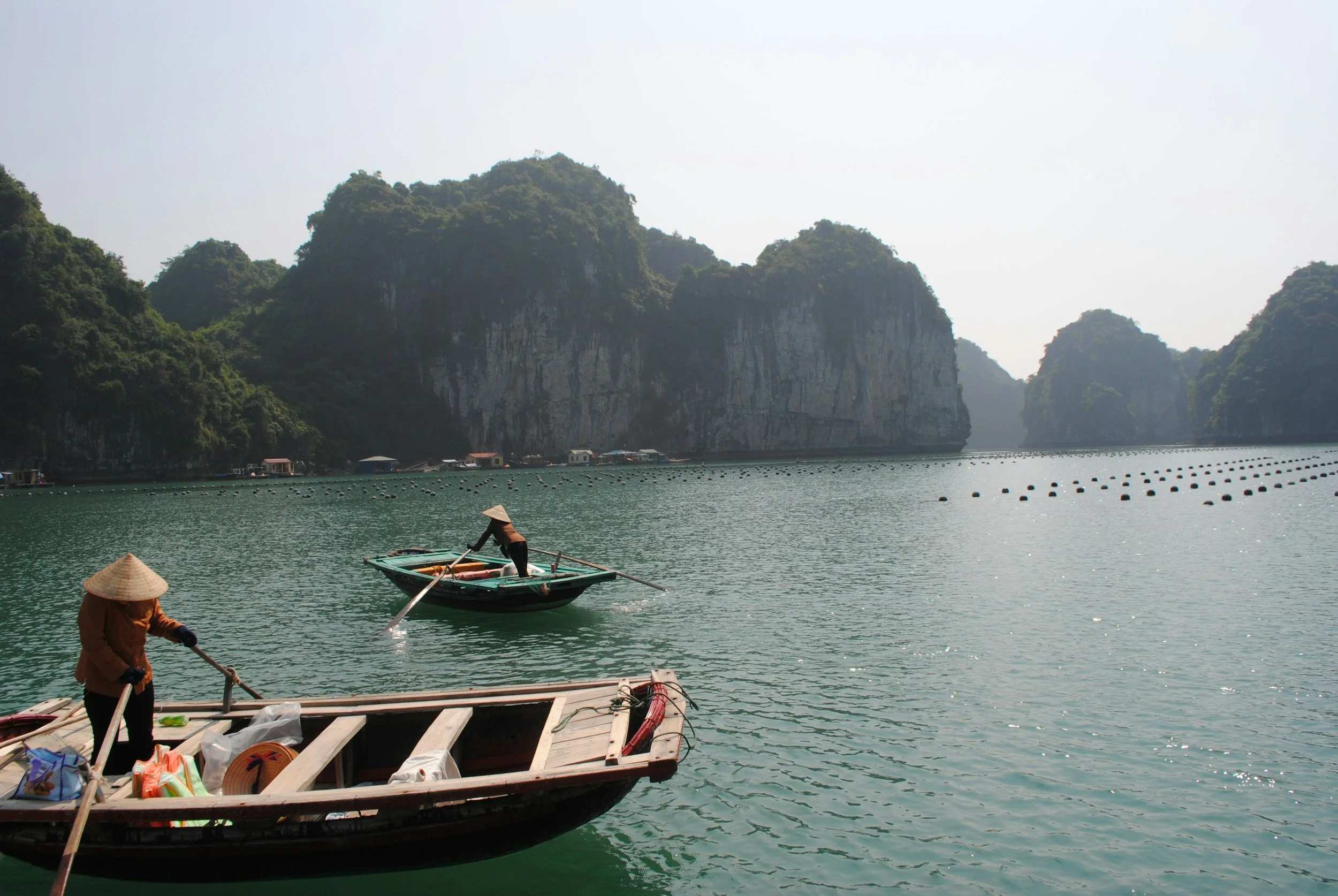 Two women in traditional conical hats rowing boats on a calm, green bay with limestone karsts in the background.