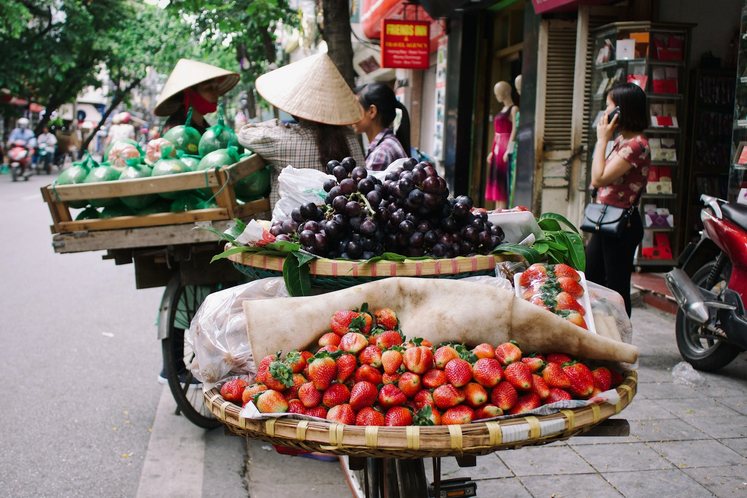 Street vendor bicycle cart with baskets of strawberries, black grapes, and watermelons on a busy sidewalk in Vietnam.
