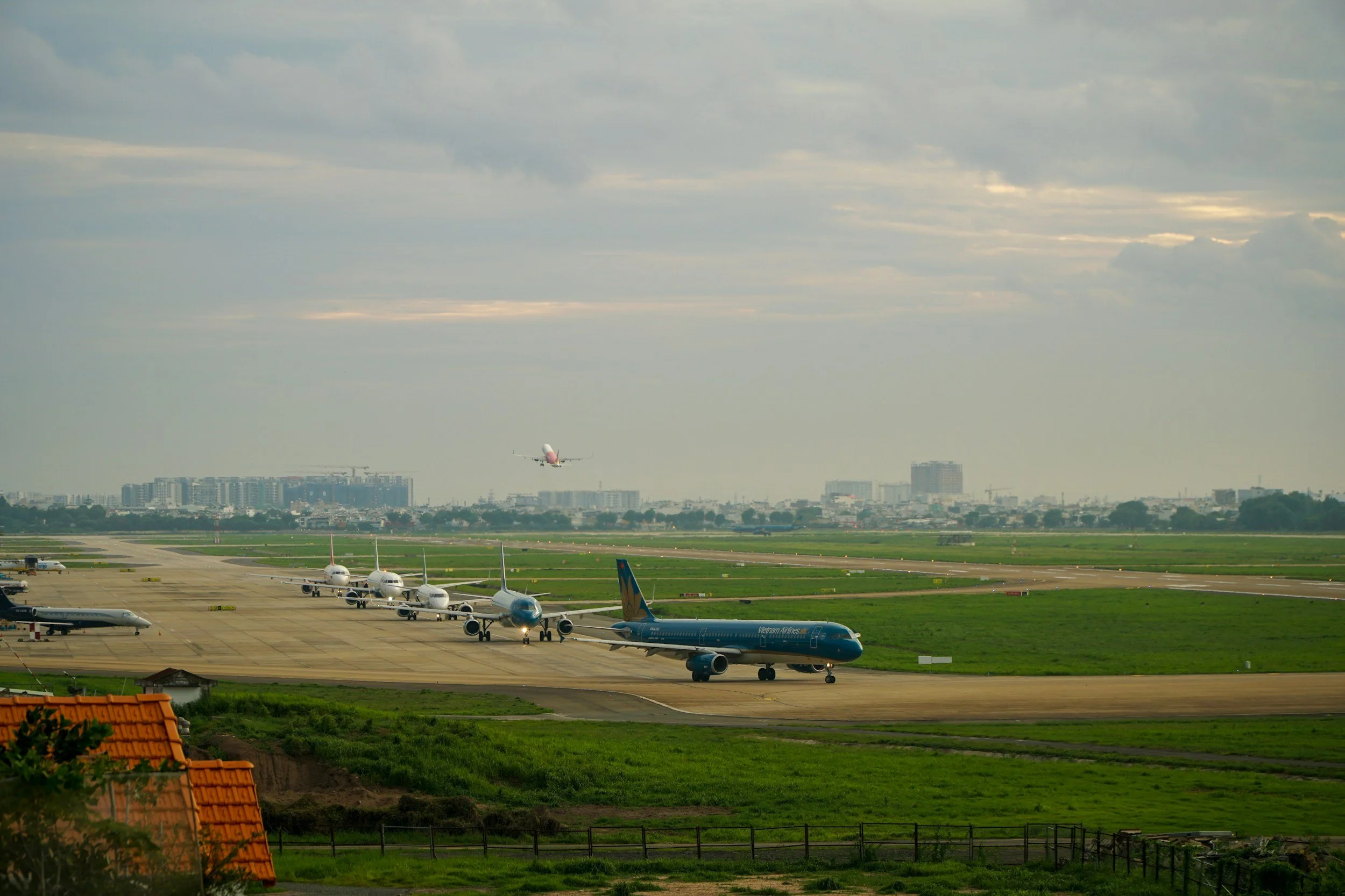 Multiple airplanes on a runway at an airport with one plane taking off in the background and city buildings in the distance under a cloudy sky.
